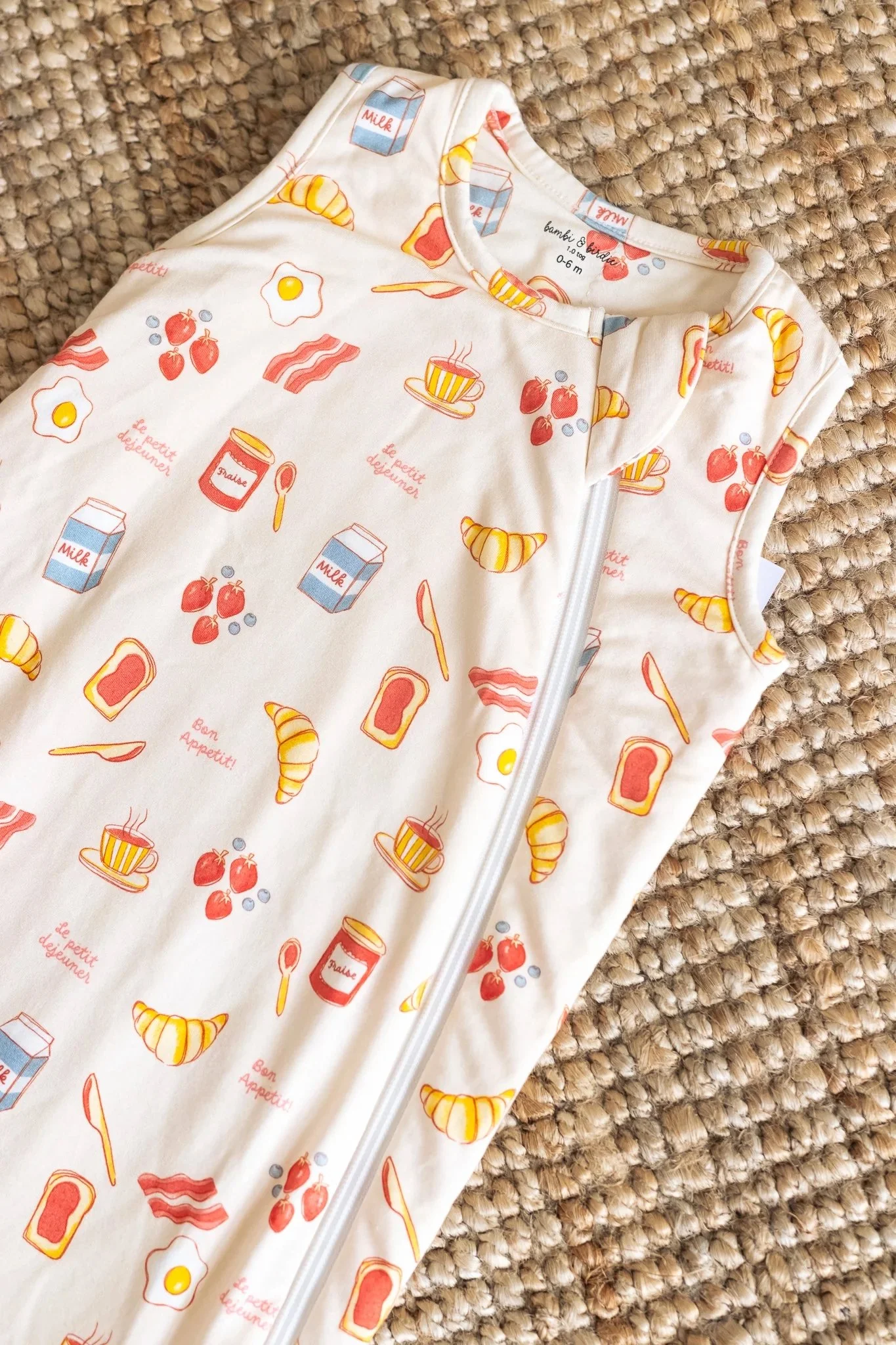 A sleeveless zipper-up baby outfit with breakfast-themed illustrations including eggs, bacon, croissants, milk cartons, and jam jars, lying on a textured woven carpet.