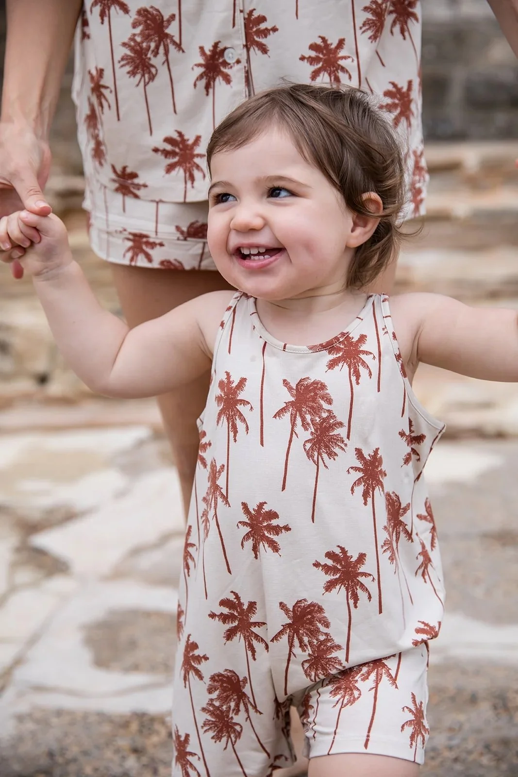 A young girl smiling and holding an adult's hand. She is wearing a sleeveless outfit with a palm tree pattern and is outdoors on a stone surface.