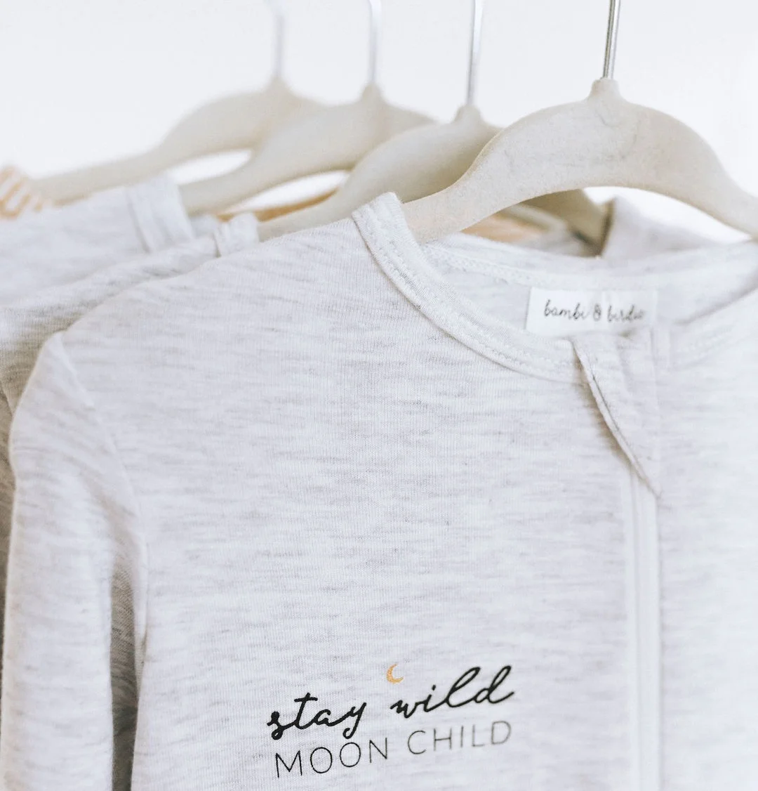 Close-up of a white baby onesie with the words "stay wild MOON CHILD" and a small moon graphic, hanging on a light-colored hanger alongside other neutral-colored baby clothes.