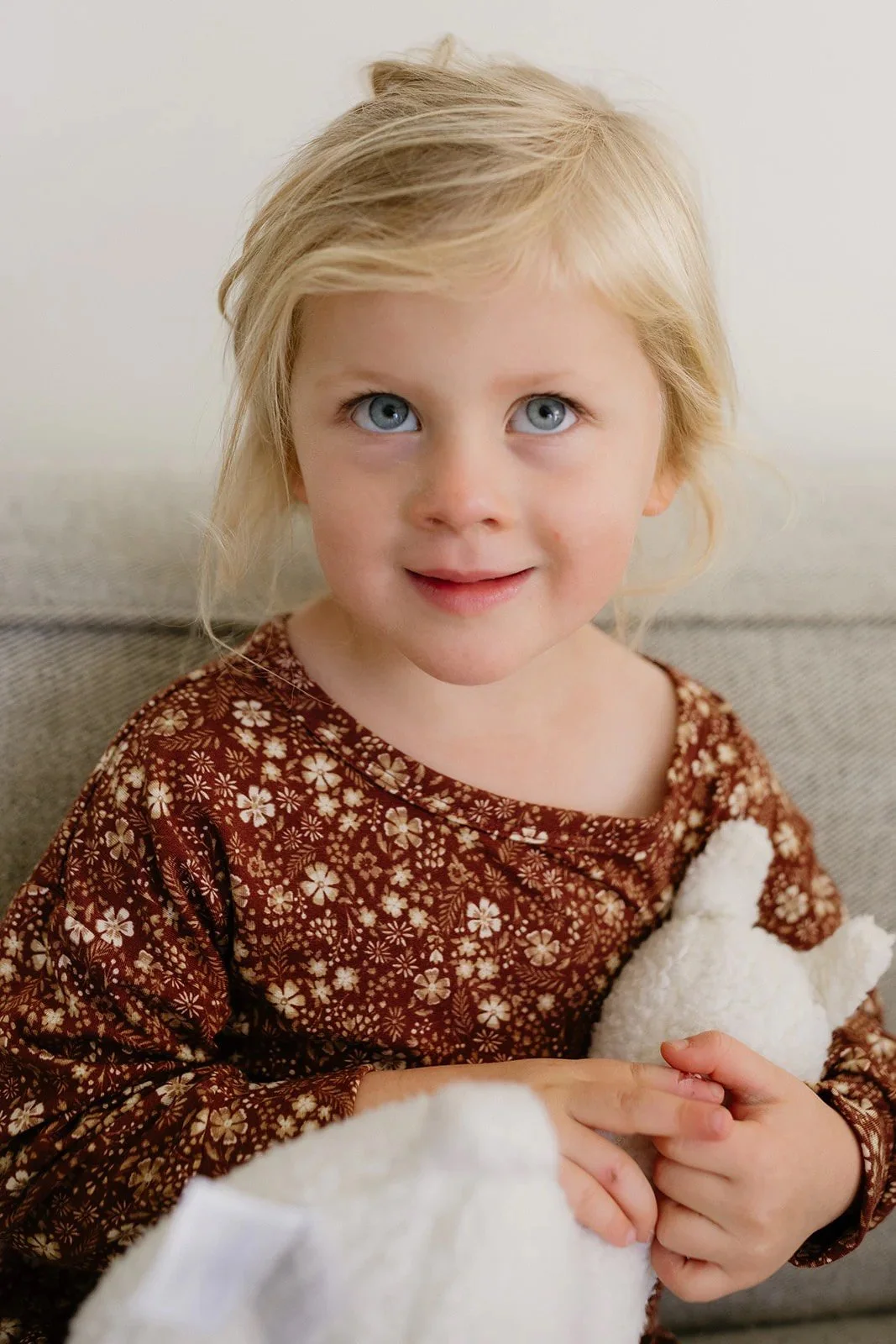 A young girl with blonde hair and blue eyes, wearing a brown floral shirt, holding a white stuffed animal, sitting on a beige couch in a light-colored room.