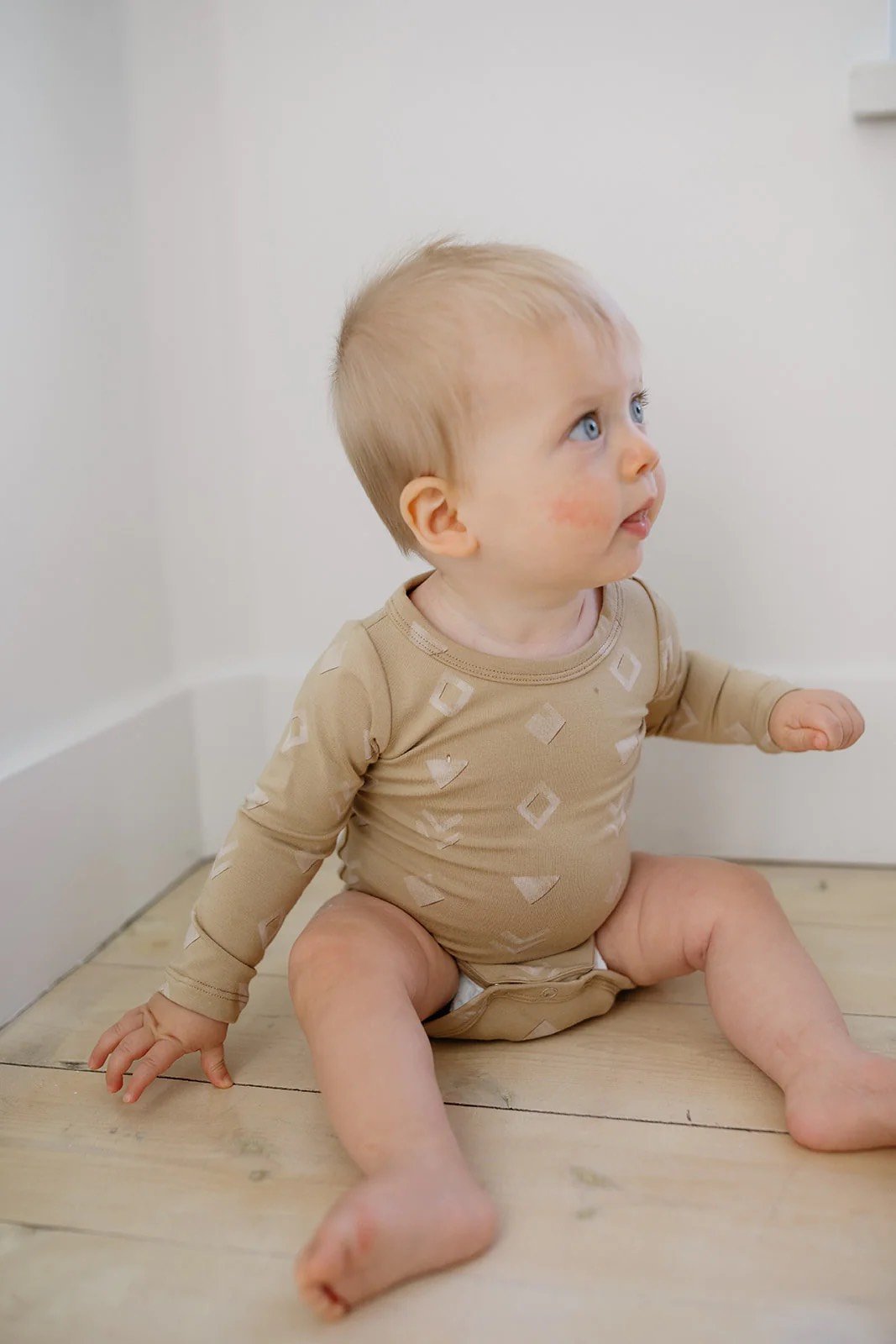 A young child with blonde hair, blue eyes, and light skin sitting on a wooden floor, looking up and to the side with a curious expression, wearing a beige long-sleeve shirt with a geometric pattern.