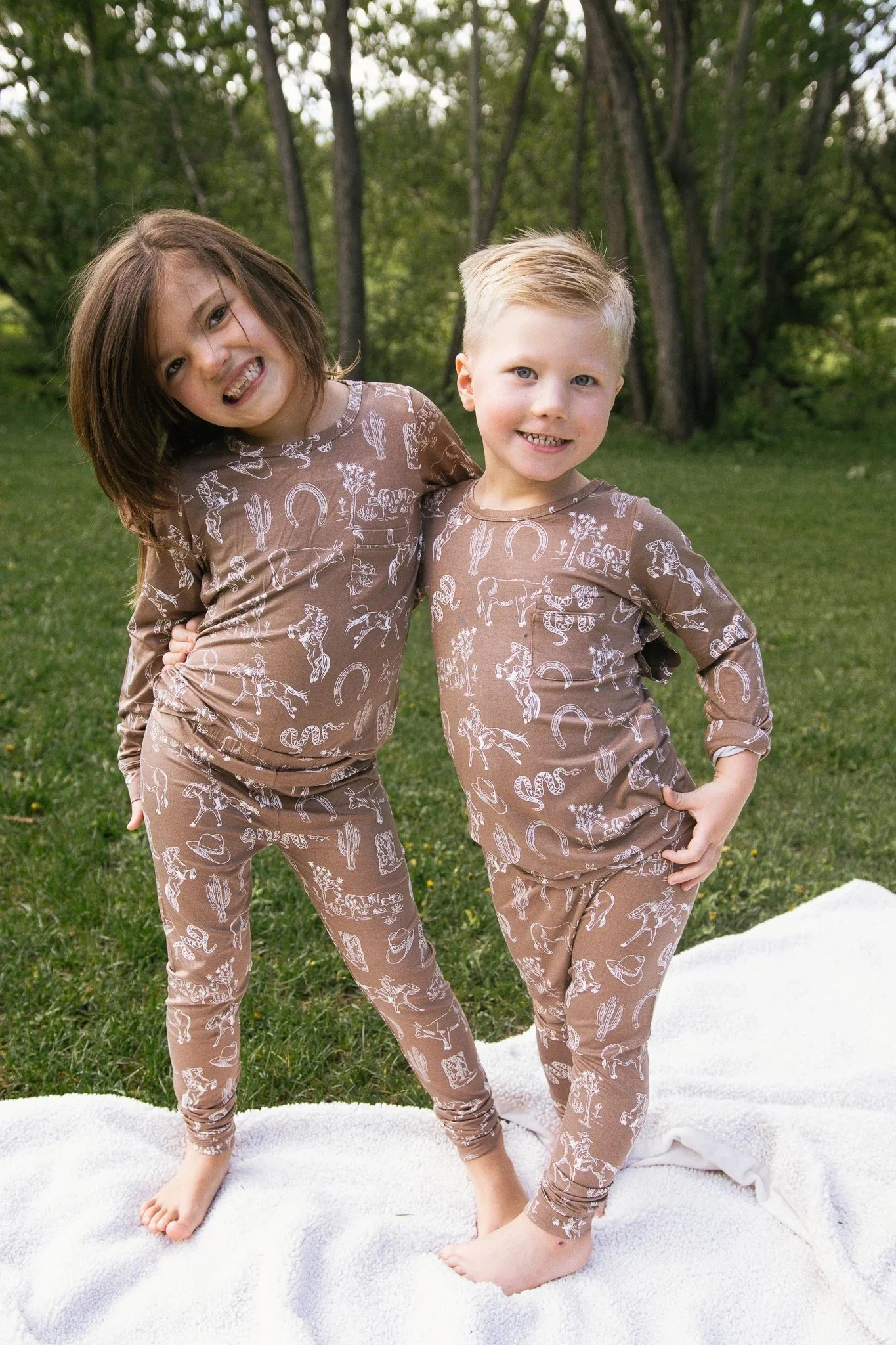 Two children standing outdoors on a white blanket, wearing matching animal-themed pajamas, smiling with arms around each other, surrounded by green grass and trees.