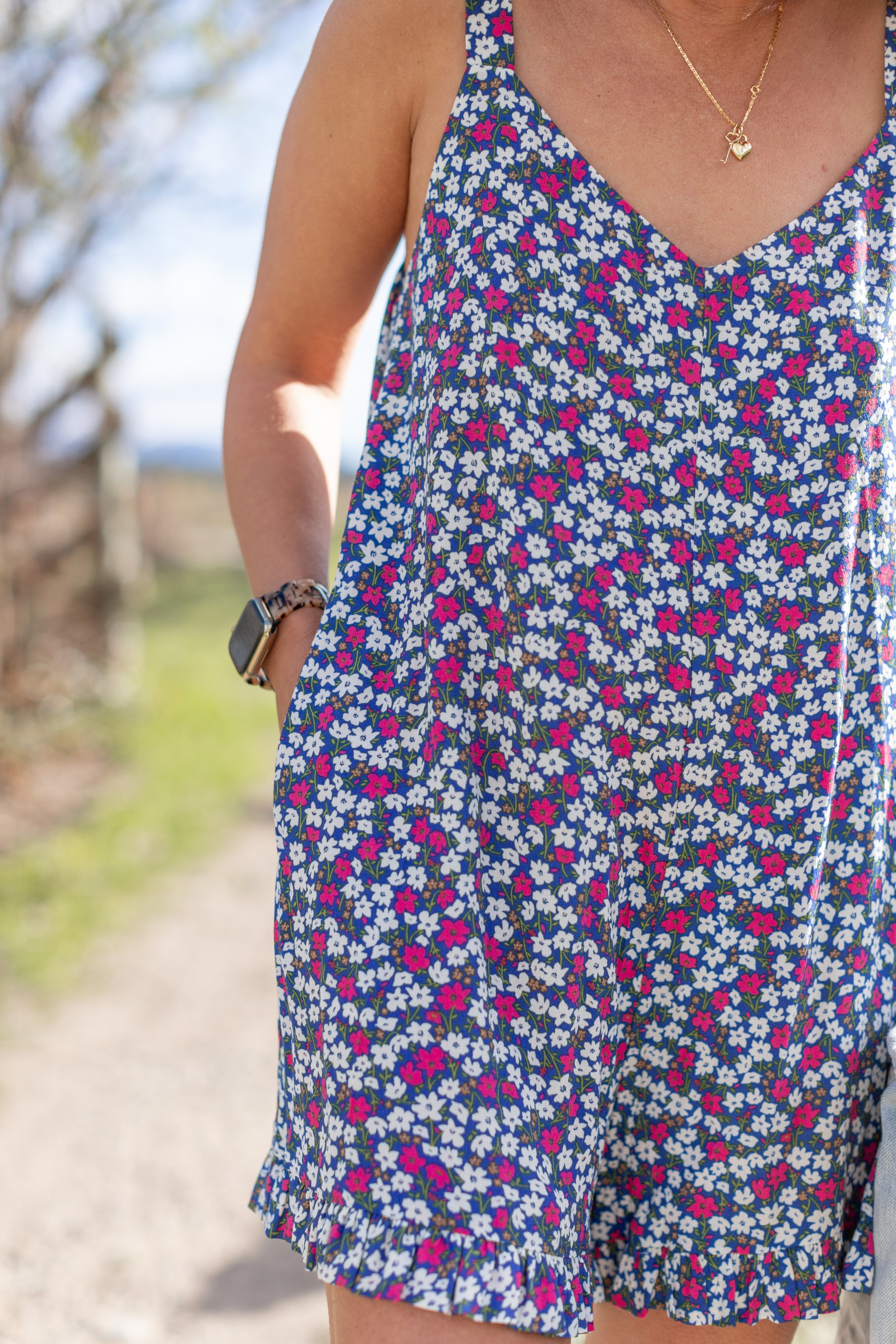 Close-up of a woman wearing a blue floral dress with pink and white flowers, a gold necklace with a heart pendant, a black watch, and a wristband, standing outdoors on a sunny day.