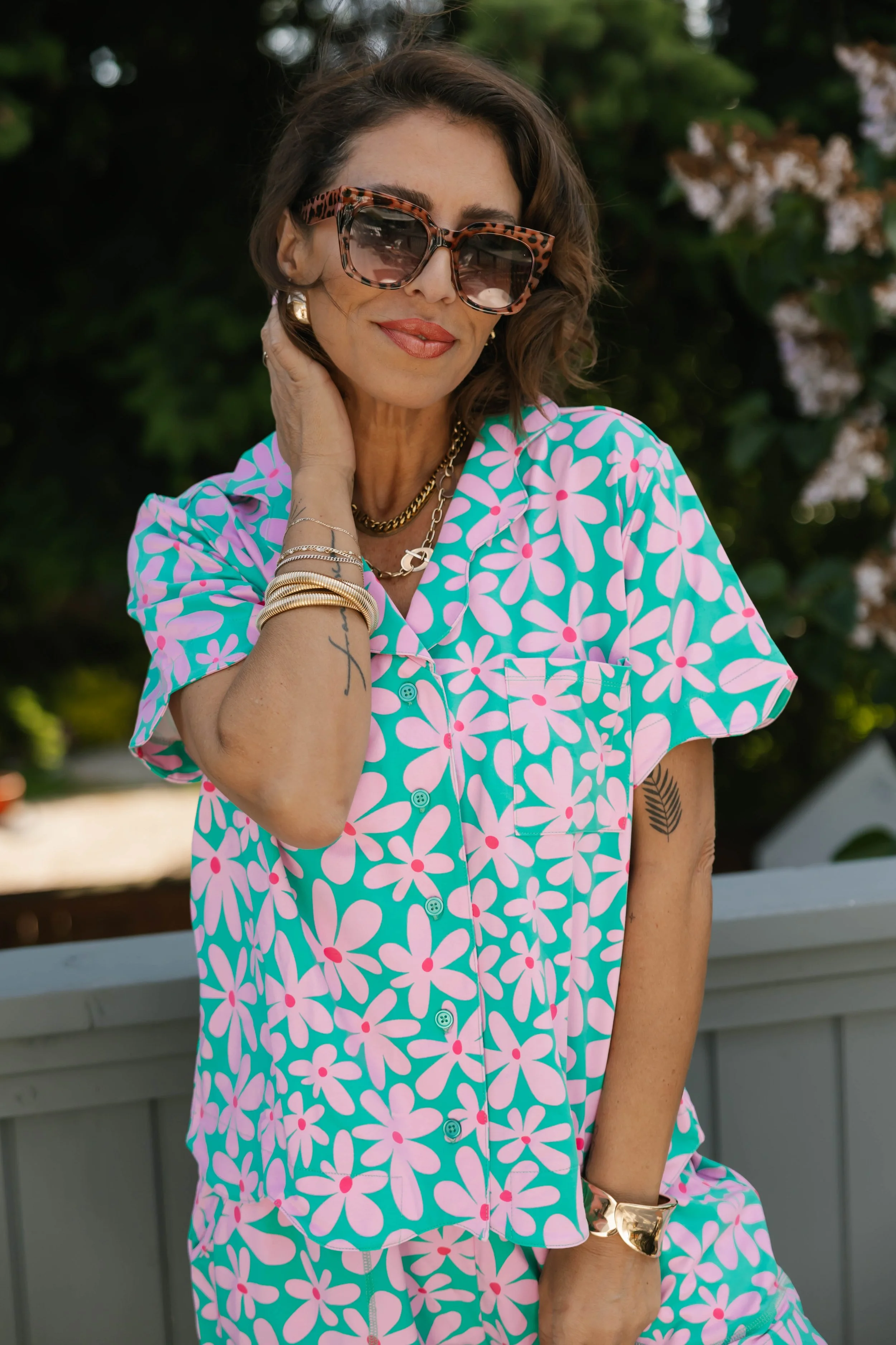 A woman wearing large, leopard-print sunglasses and colorful, floral-patterned shirt, standing outdoors near a gray railing with greenery and flowers in the background.
