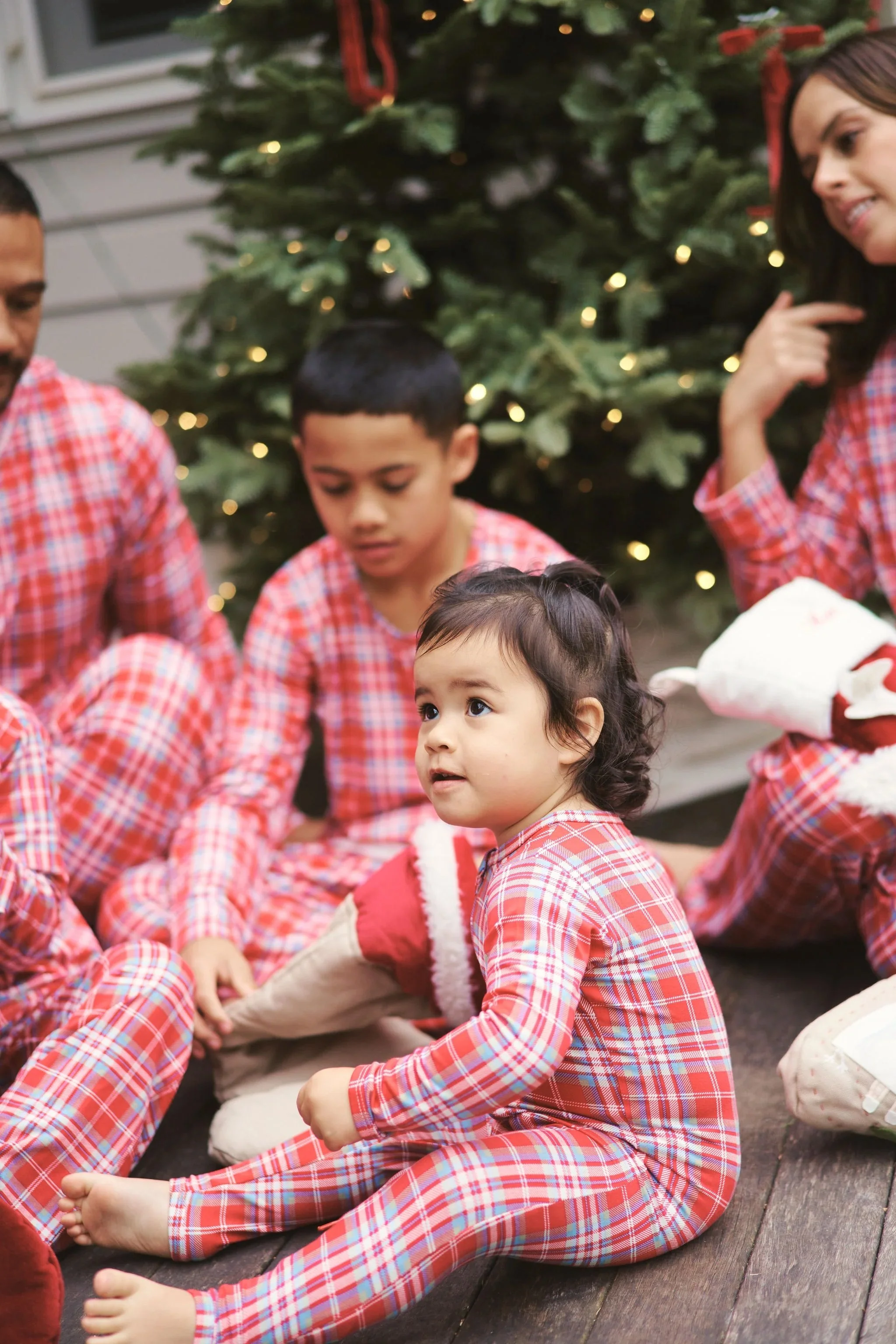 A family celebrating Christmas indoors, wearing matching red and white plaid pajamas, sitting on the floor in front of a decorated Christmas tree with lights.