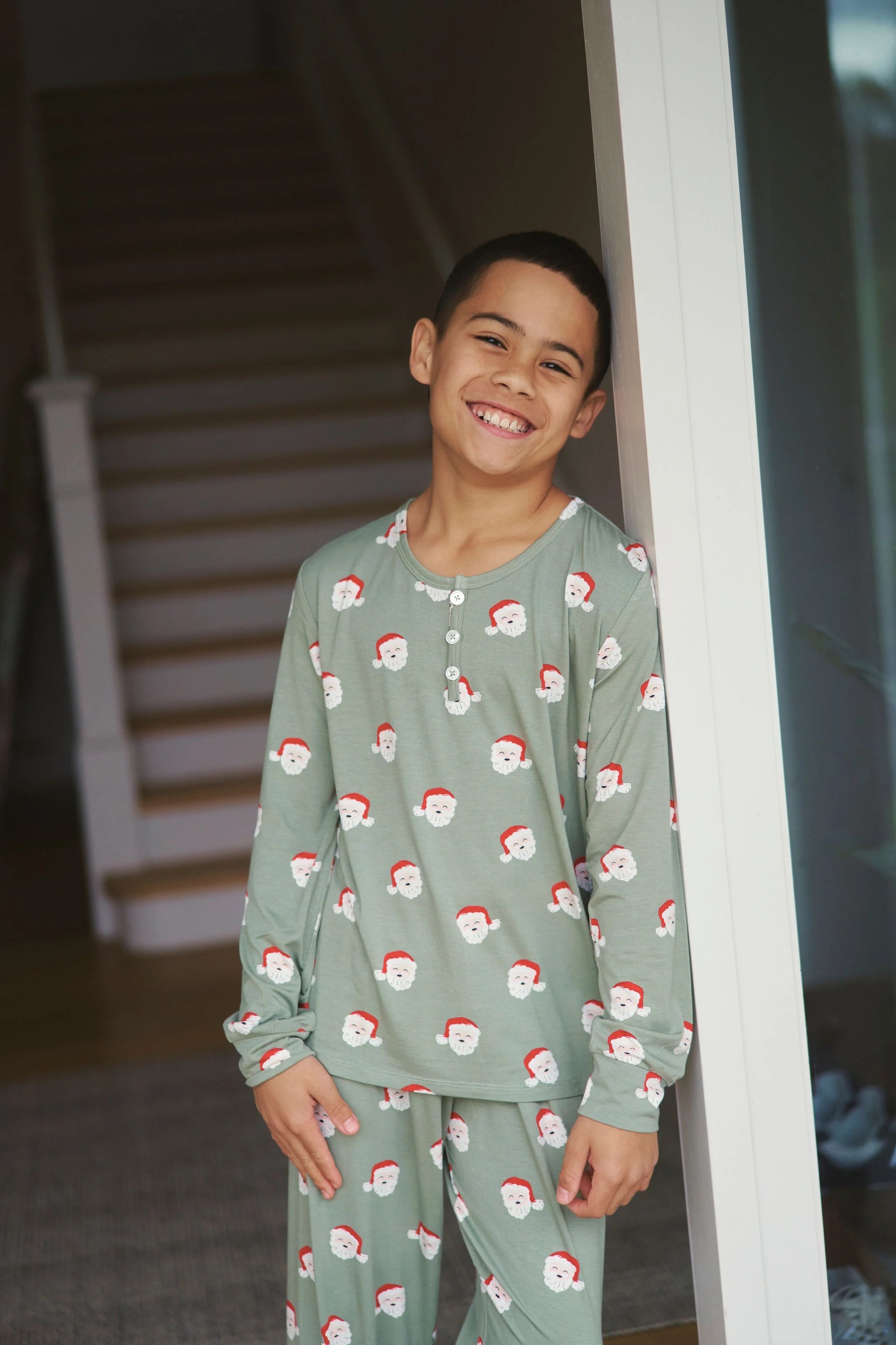 Smiling boy wearing green pajamas with Santa Claus faces, leaning against a doorway near stairs in a home.