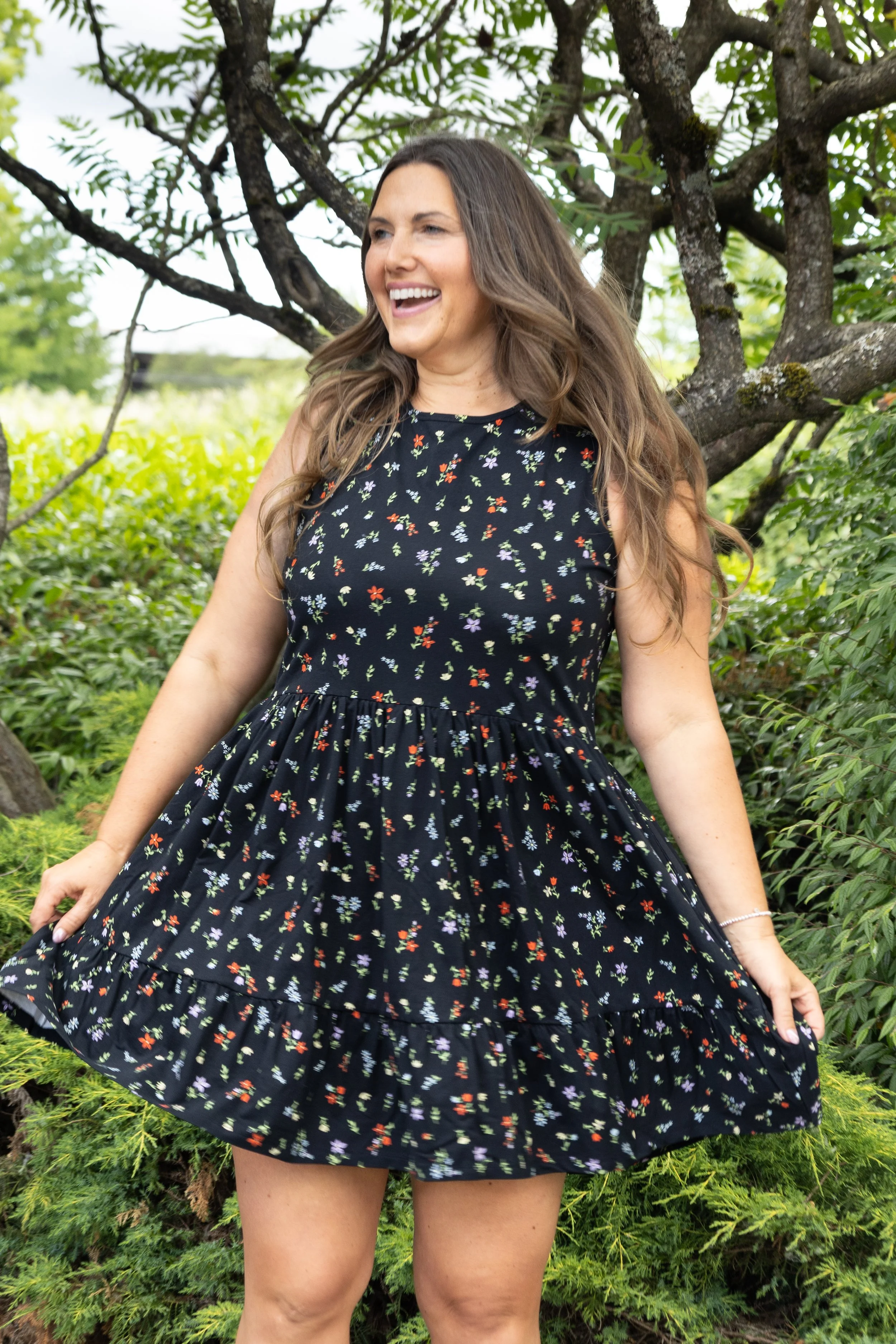 A smiling woman with long, wavy brown hair wearing a black dress with a colorful floral pattern, standing outdoors near greenery and a tree.