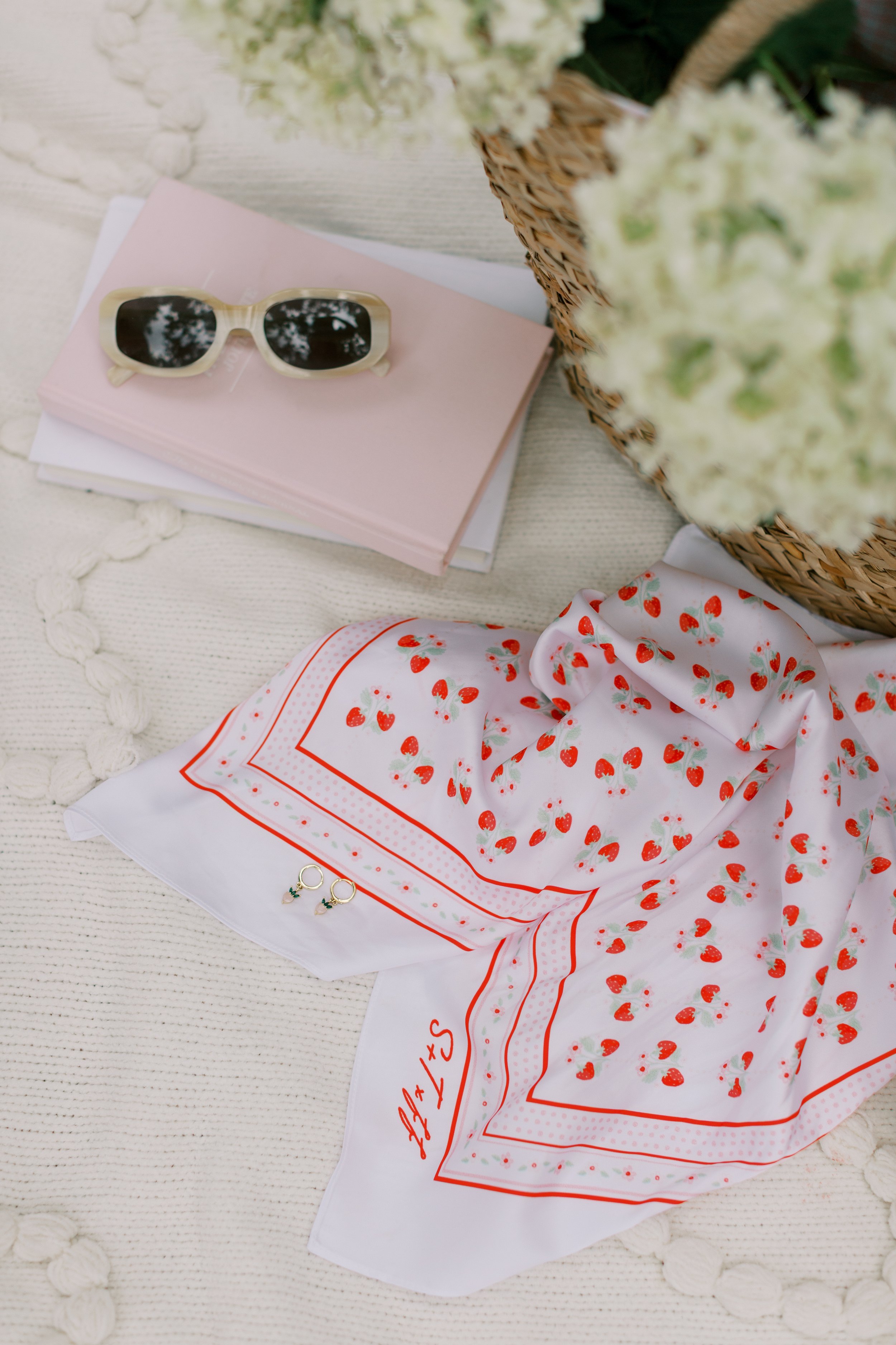 A flat lay of sunglasses on a pink notebook, a white scarf with a strawberry pattern, a pair of earrings, and a wicker basket filled with white flowers, all on a textured white surface.