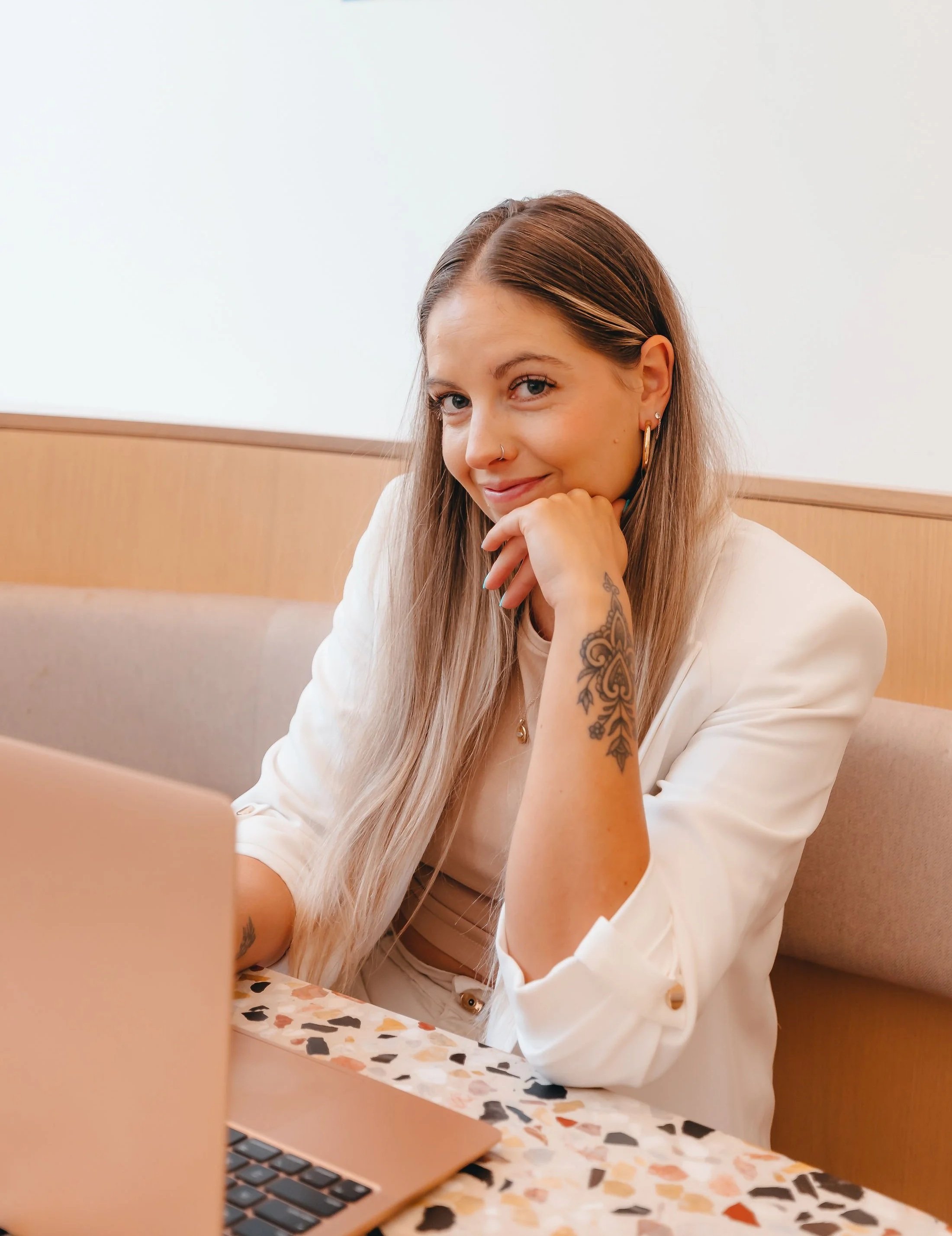 Young woman with long, straight hair, earrings, and a nose ring, smiling and resting her chin on her hand, sitting at a terrazzo table with a laptop and a beige booth.