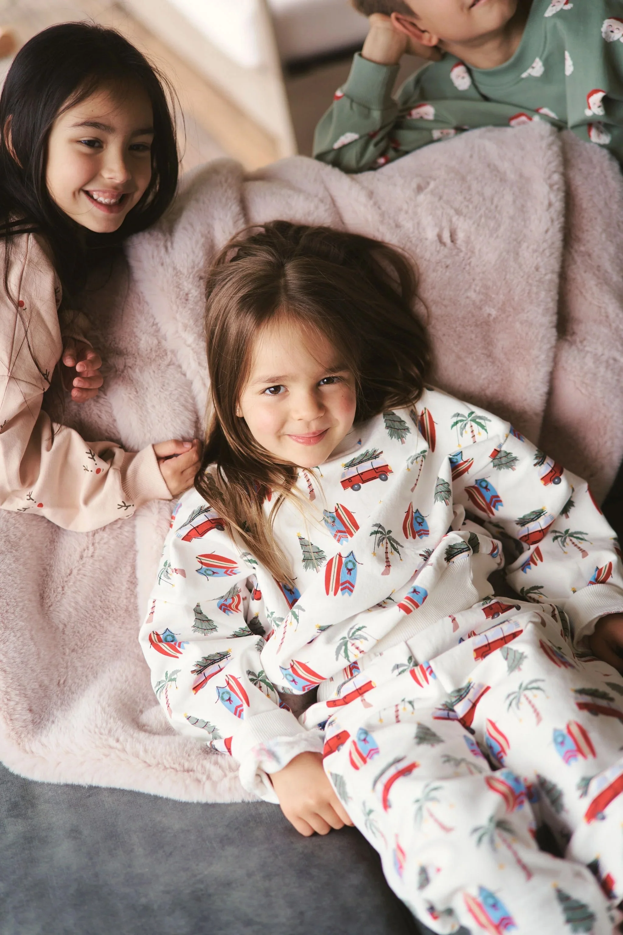 Three children, two girls and one boy, are lying down on a soft beige blanket. The girl with dark hair is smiling at the camera, while the girl with brown hair is looking directly with a slight smile, dressed in festive pajamas with holiday motifs. The boy, wearing Christmas-themed pajamas, is lying partly out of the frame, resting his head on his hand.