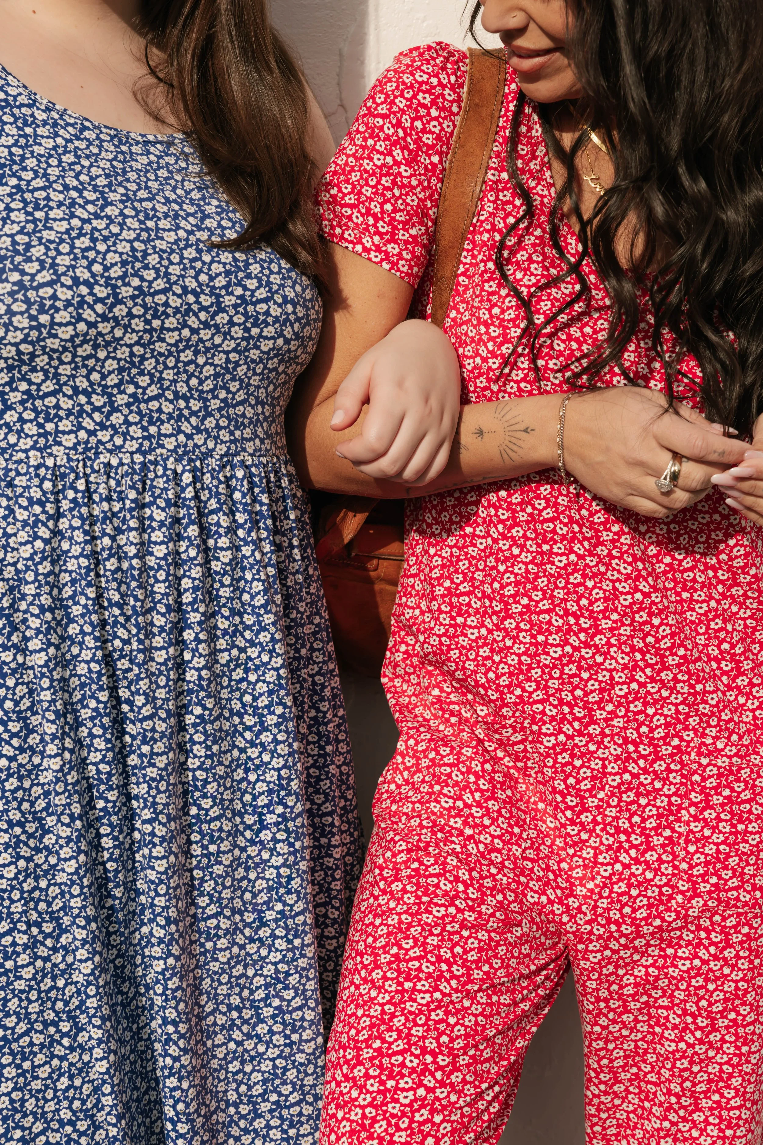 Two women standing close, holding hands. One woman wears a blue floral dress, the other wears a red floral dress. The woman in red has long dark hair, jewelry, and a tattoo on her wrist.