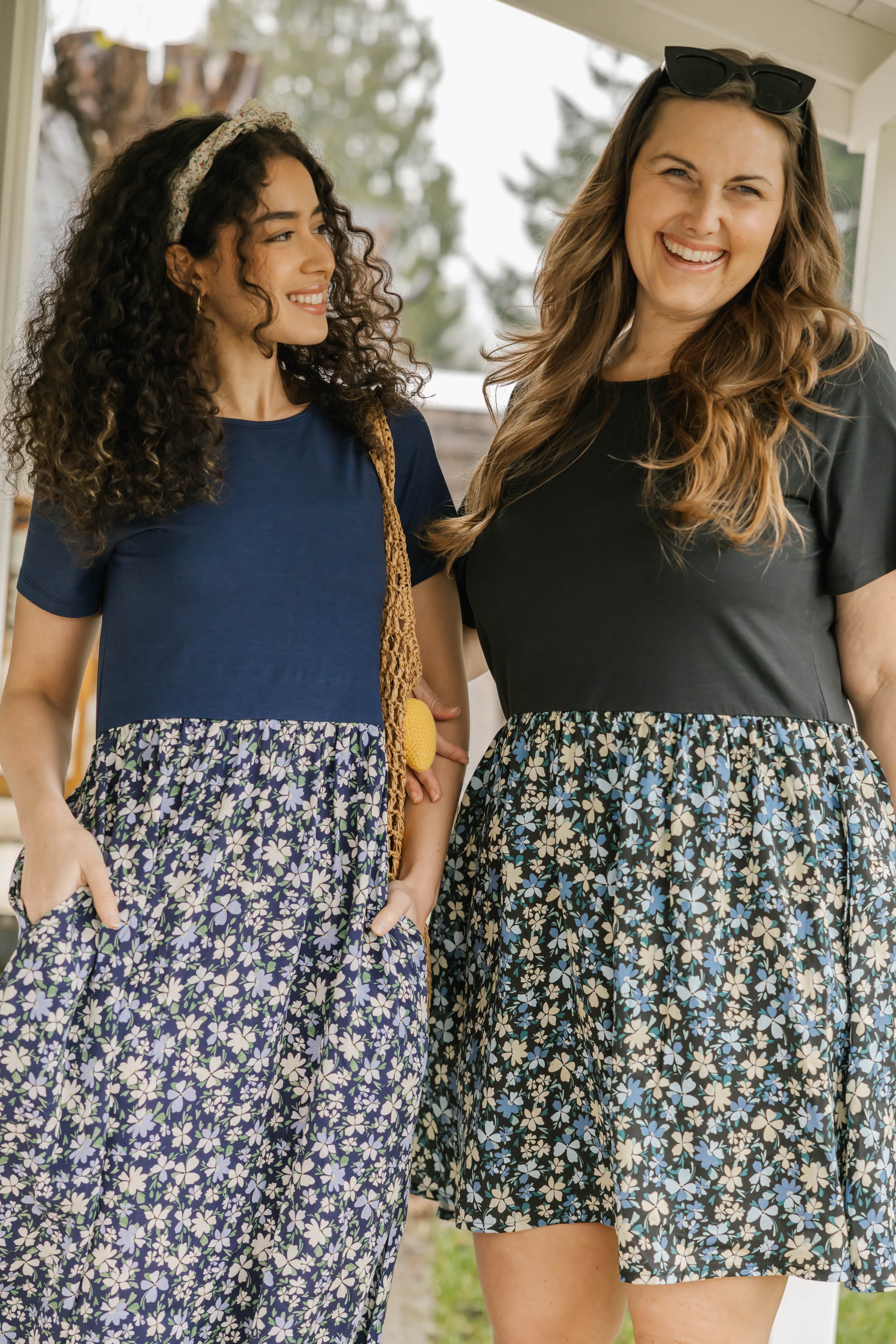 Two women standing outside, smiling and looking at each other. One woman has dark curly hair, wearing a navy blue top and floral skirt, with a headband. The other woman has light brown wavy hair, wearing sunglasses on her head, a black top, and a flo