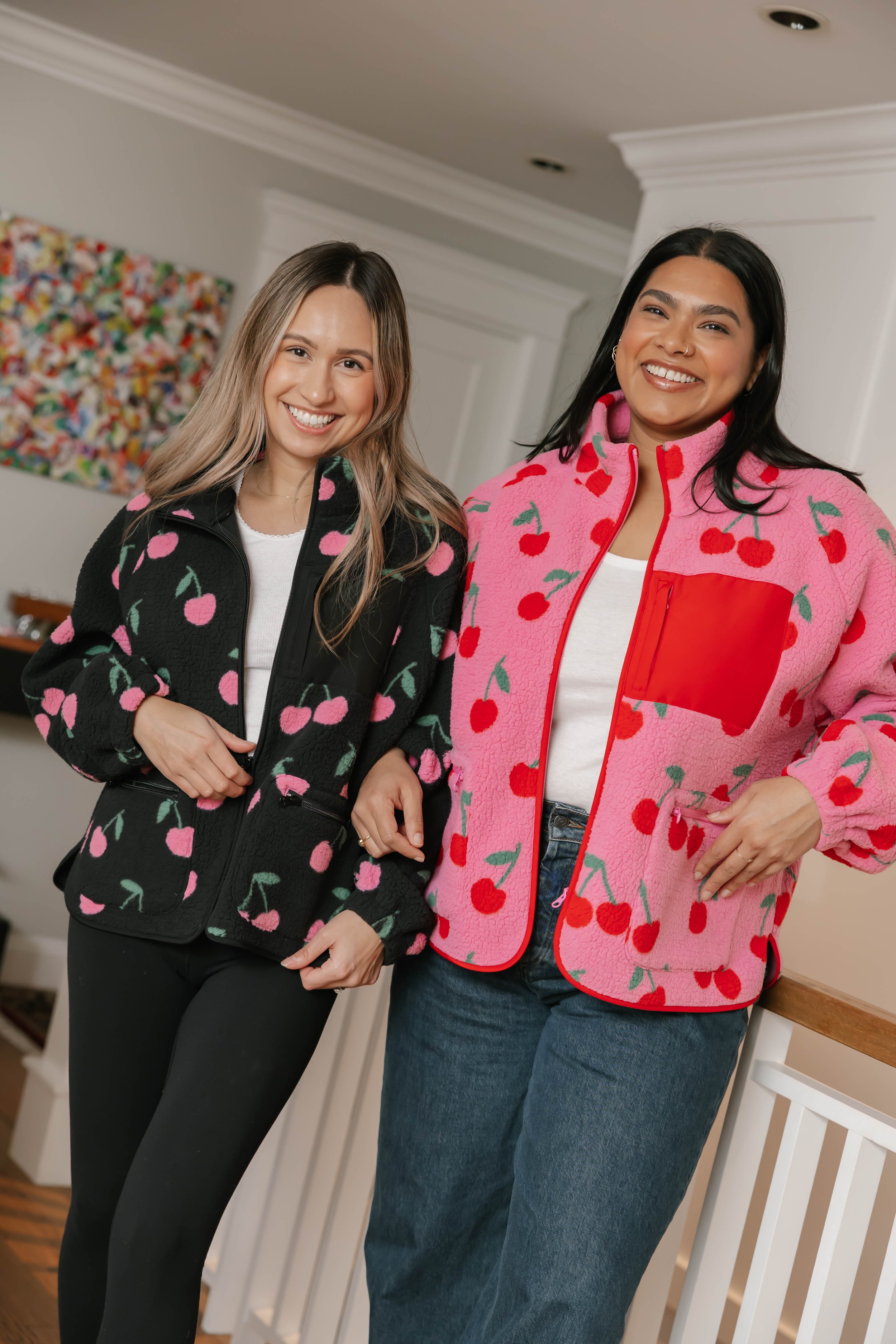 Two women standing indoors, smiling, wearing cherry-patterned fleece jackets, one black and one pink, with a colorful painting on the wall behind them.