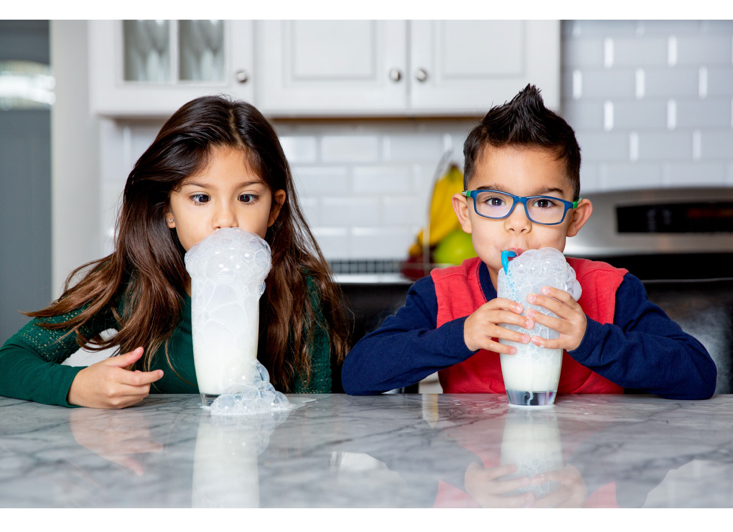  Two kids sit at a kitchen counter blowing bubbles into their glasses of milk. 