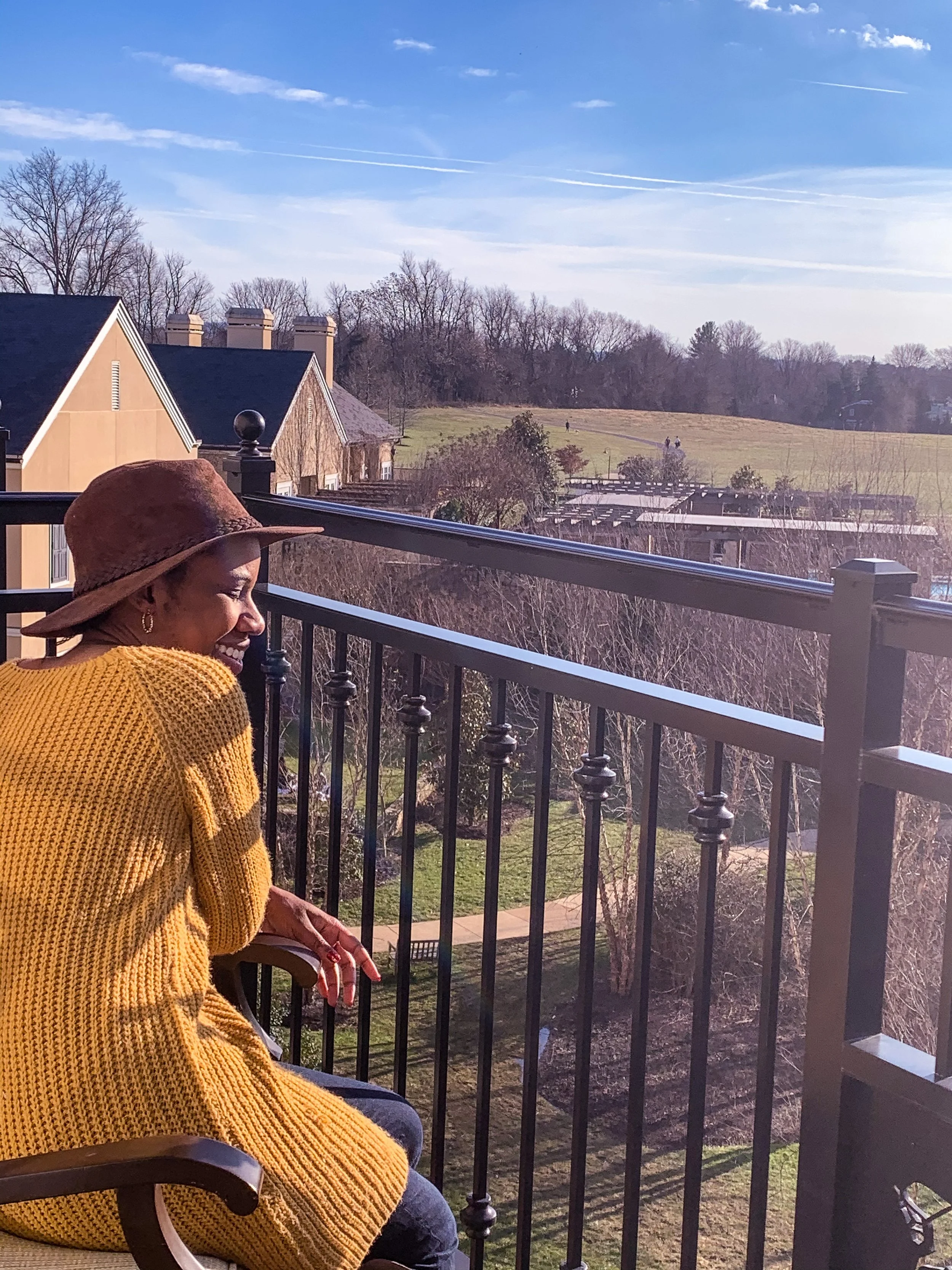Woman in a yellow sweater and brown hat sitting on a balcony with a view of trees, houses, and an open field under a blue sky.