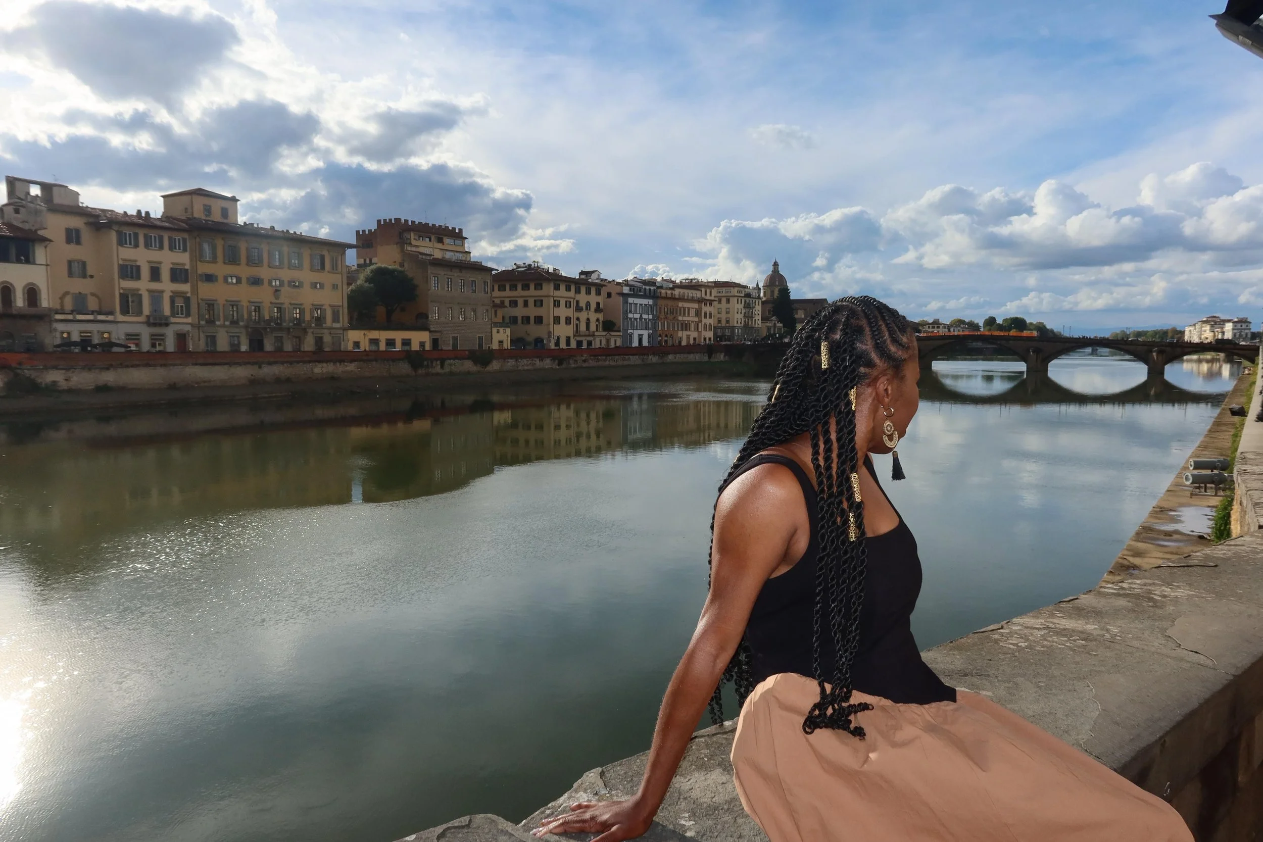 A woman with braided hair and earrings sitting on a stone ledge by a river in a city with historic buildings, a bridge, and cloudy sky