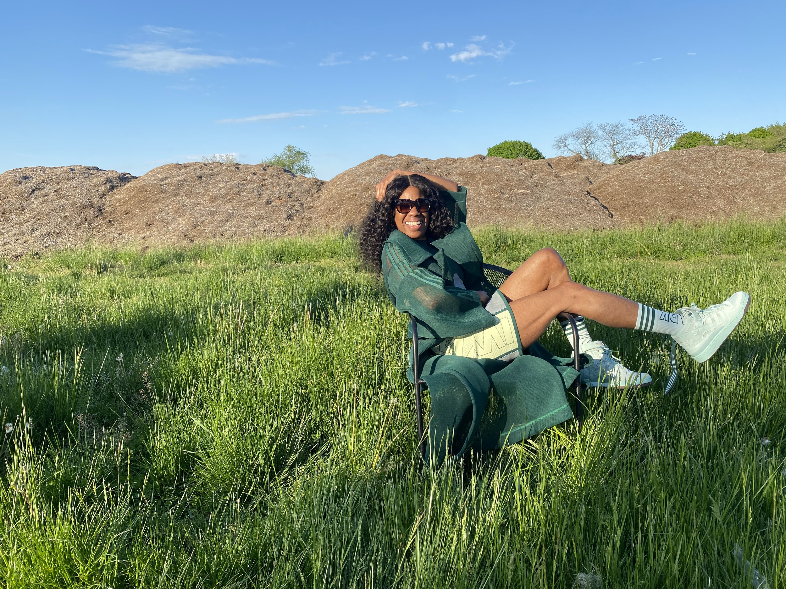A woman with curly black hair, sunglasses, a green jacket, and white sneakers, sitting on a green camping chair in a grassy field, smiling during a sunny day with blue sky and rocks in the background.