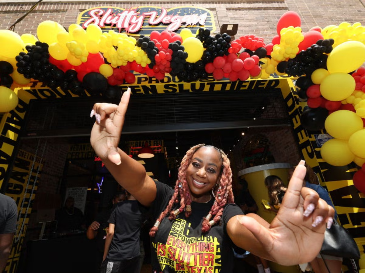 Woman with pink braids smiling and pointing at the camera, standing under an arch decorated with red, yellow, and black balloons at a vegan restaurant called Slutty Vegan.
