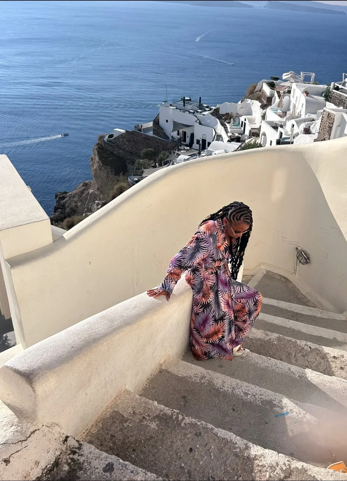 A woman in a colorful tropical print outfit sitting on a curved white staircase overlooking the sea and white buildings of a coastal town, with boats in the water.