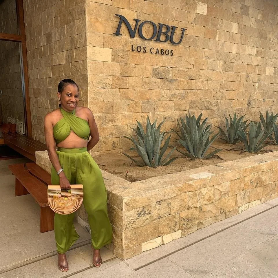 A woman in a green outfit standing next to a stone planter with agave plants, in front of a wall with the sign 'NOBU LOS CABOS'.