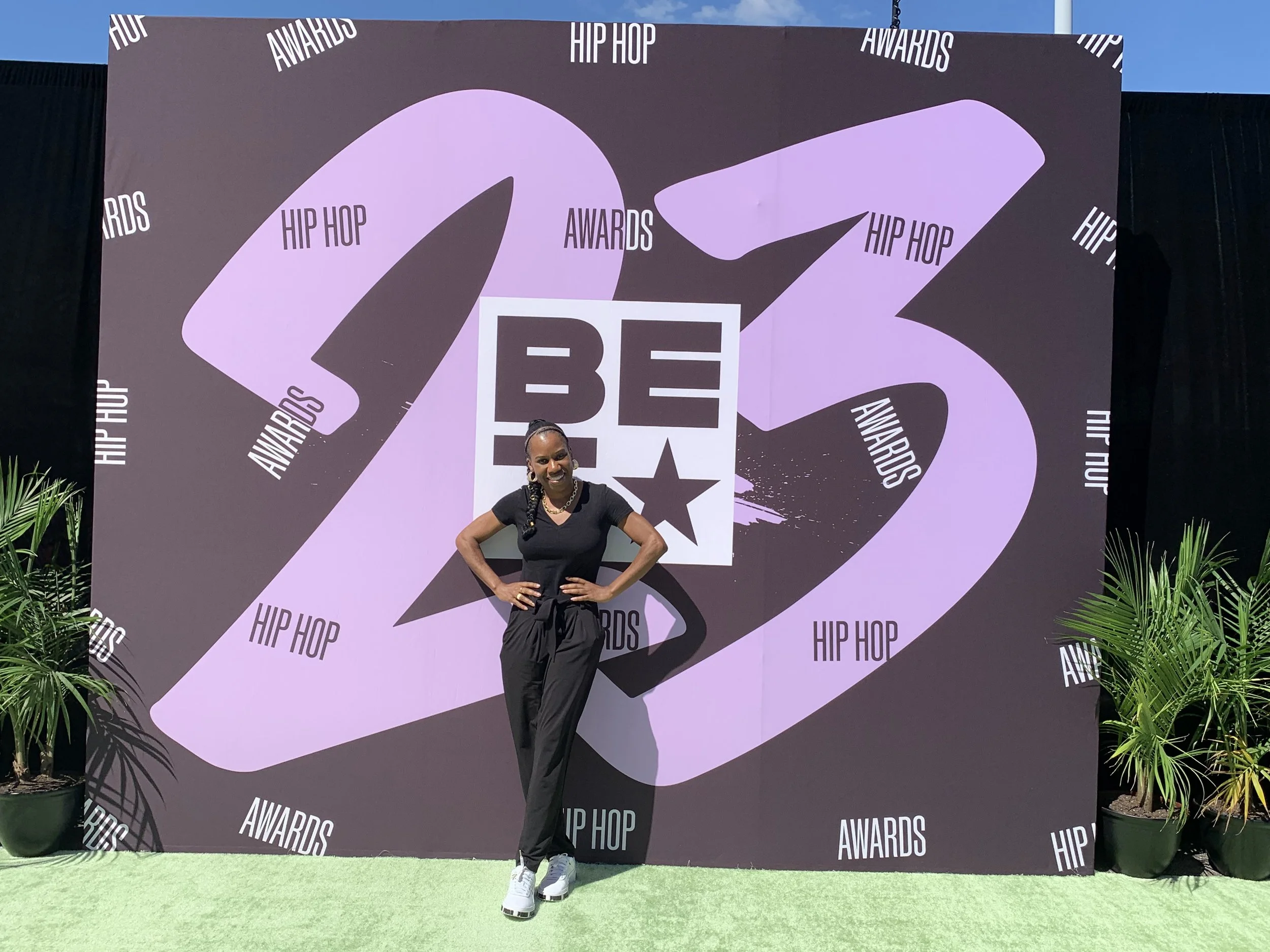 A woman smiling stands in front of a large, purple and black backdrop with the words 'HIP HOP AWARDS' and the numbers '23'. She poses with her hands on her hips and is dressed in a black T-shirt, black pants, and white sneakers, with potted plants on either side of her.