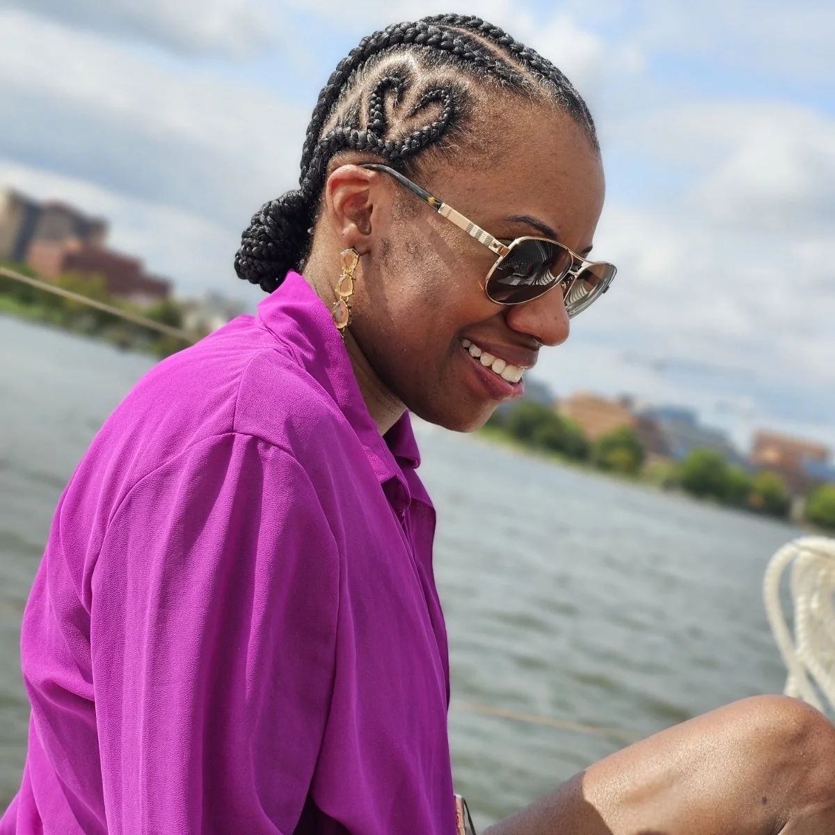 A woman with braided hair, wearing sunglasses, gold earrings, and a purple shirt, smiling outdoors near a body of water with a cityscape in the background.