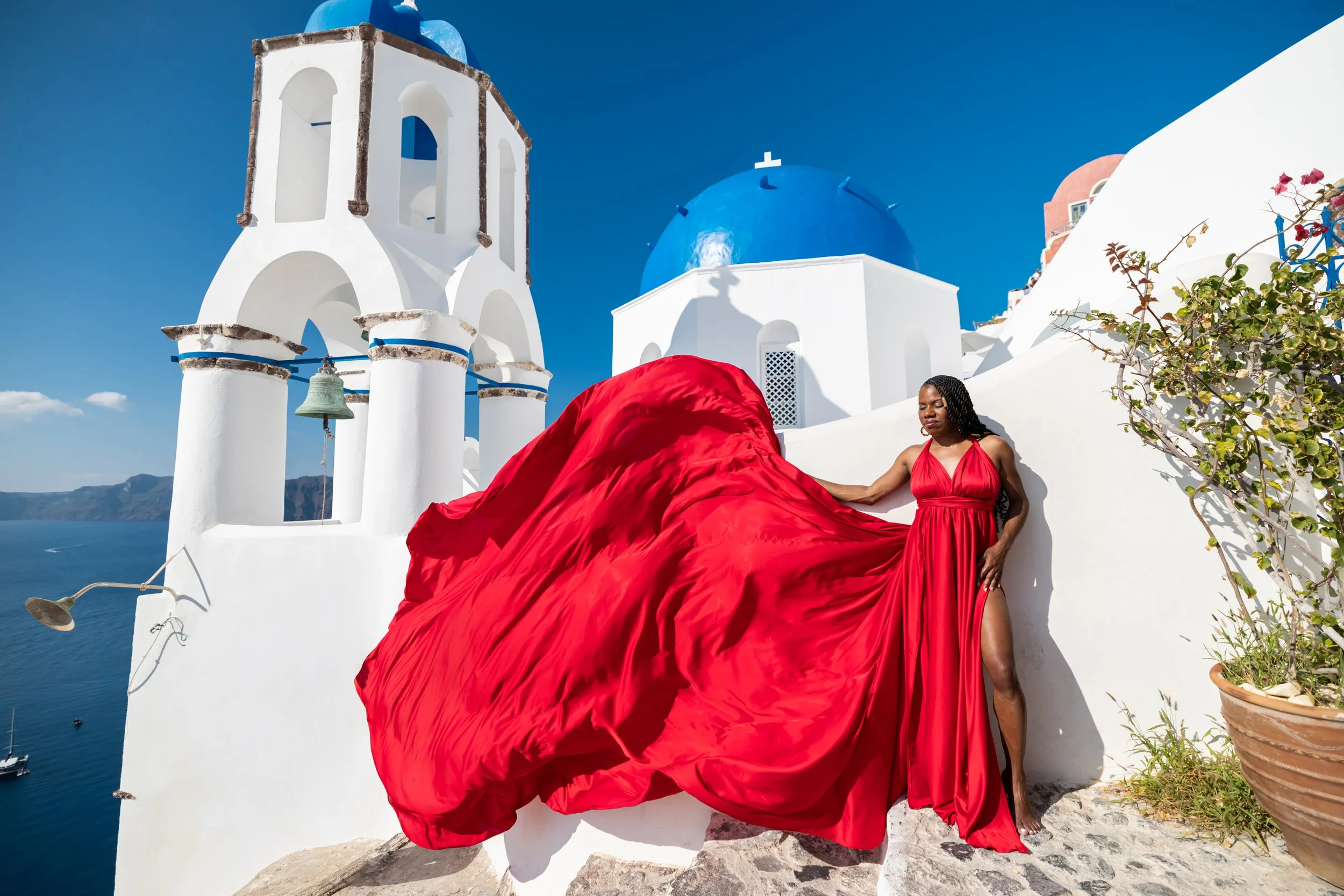 A woman in a long red gown standing against a white wall with blue domed buildings and a bell tower in Santorini, Greece, during daytime.