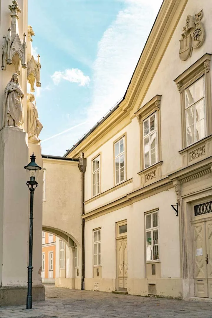 Quiet street next to Pfarrkirche Hietzing — archway leading into a lane past a house with a coat of arms in the roof section, Vienna