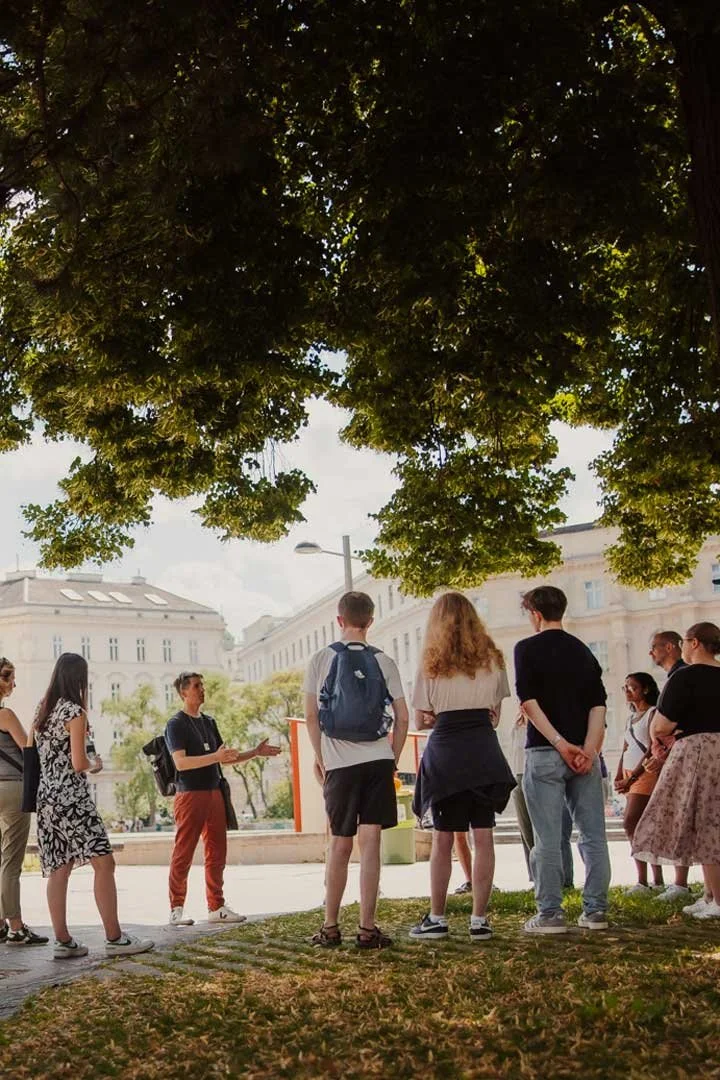 Walking tour group under a lush green tree listening to stories of imperial scandals in Hietzing, Vienna