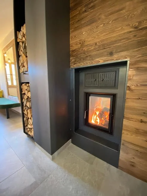 Ein moderner Kamin mit brennendem Feuer vor einer Holzverkleidung, daneben ein Holzstapel und ein Sitzbereich mit grünem Stuhl im Hintergrund.