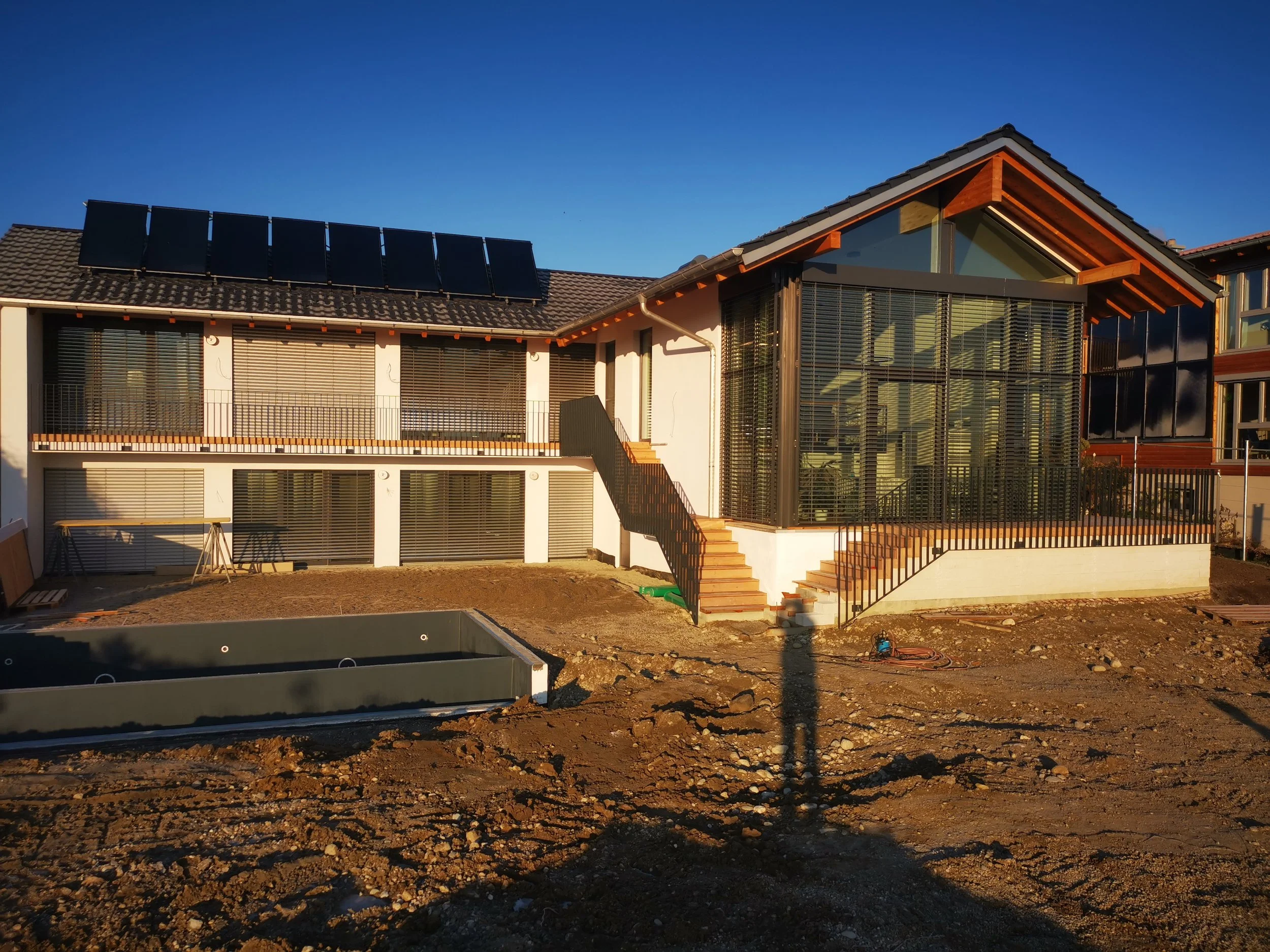 Ein modernes Haus unter Bau, mit Sonnenkollektoren auf dem Dach, großen Fenstern und einer Terrasse mit Stufen, umgeben von Erde und Baumaterialien.