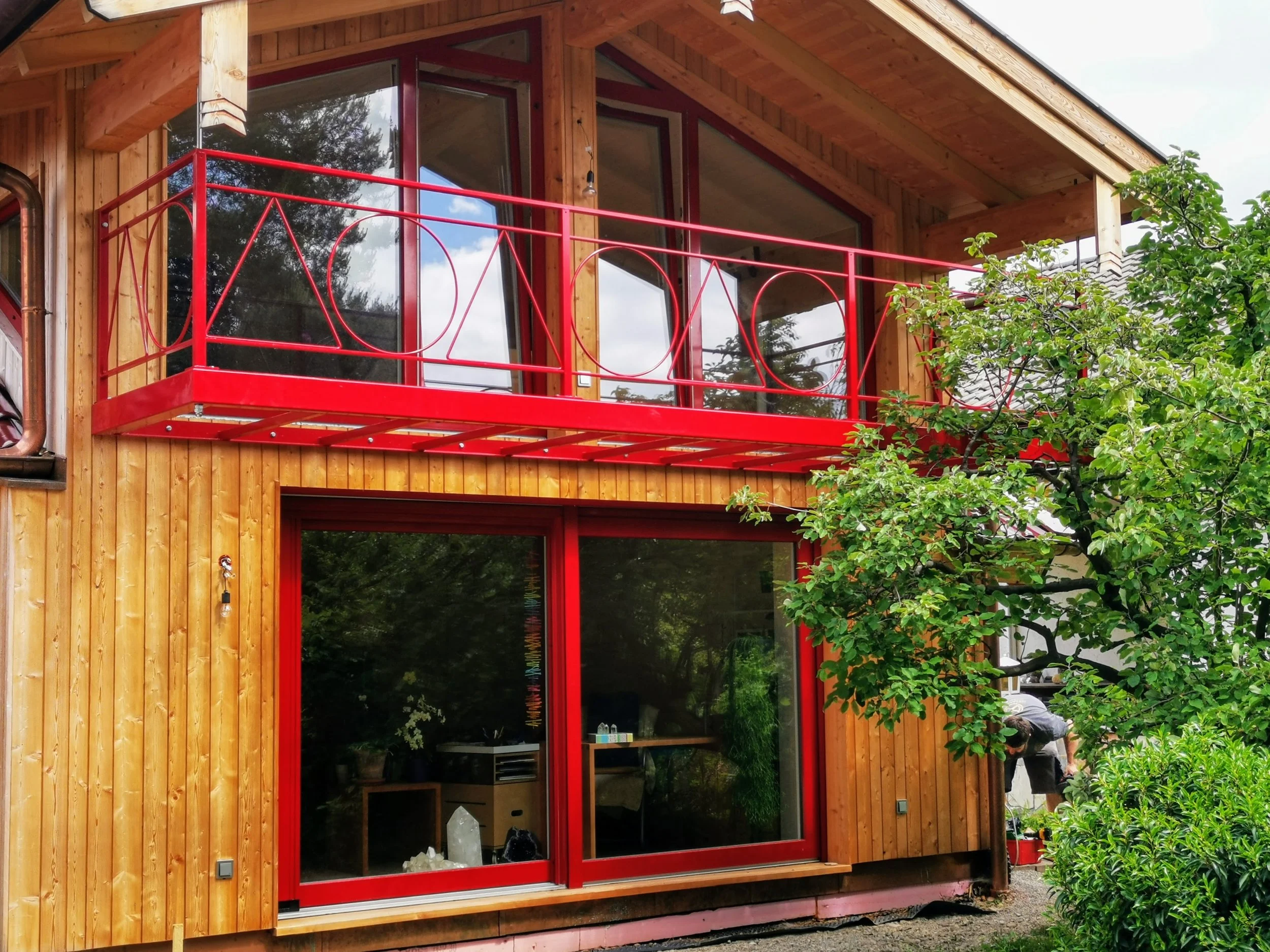 Holzhaus mit roter Balkontür und großen Fenstern, umgeben von grünen Bäumen und Sträuchern.
