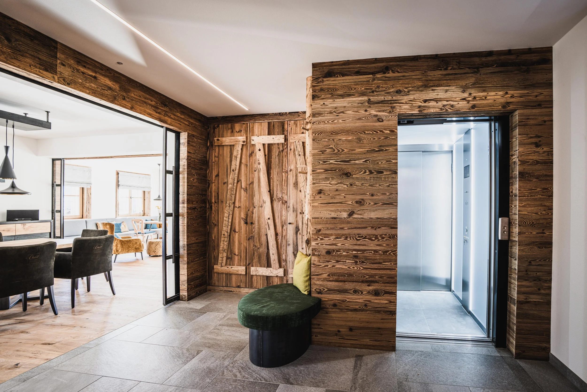 Innenaufnahme eines Gebäudes mit Holzverkleidung, einem Sessel mit grünem Bezug, einem Aufzug und einem Raum mit Esstisch und Stühlen im Hintergrund.