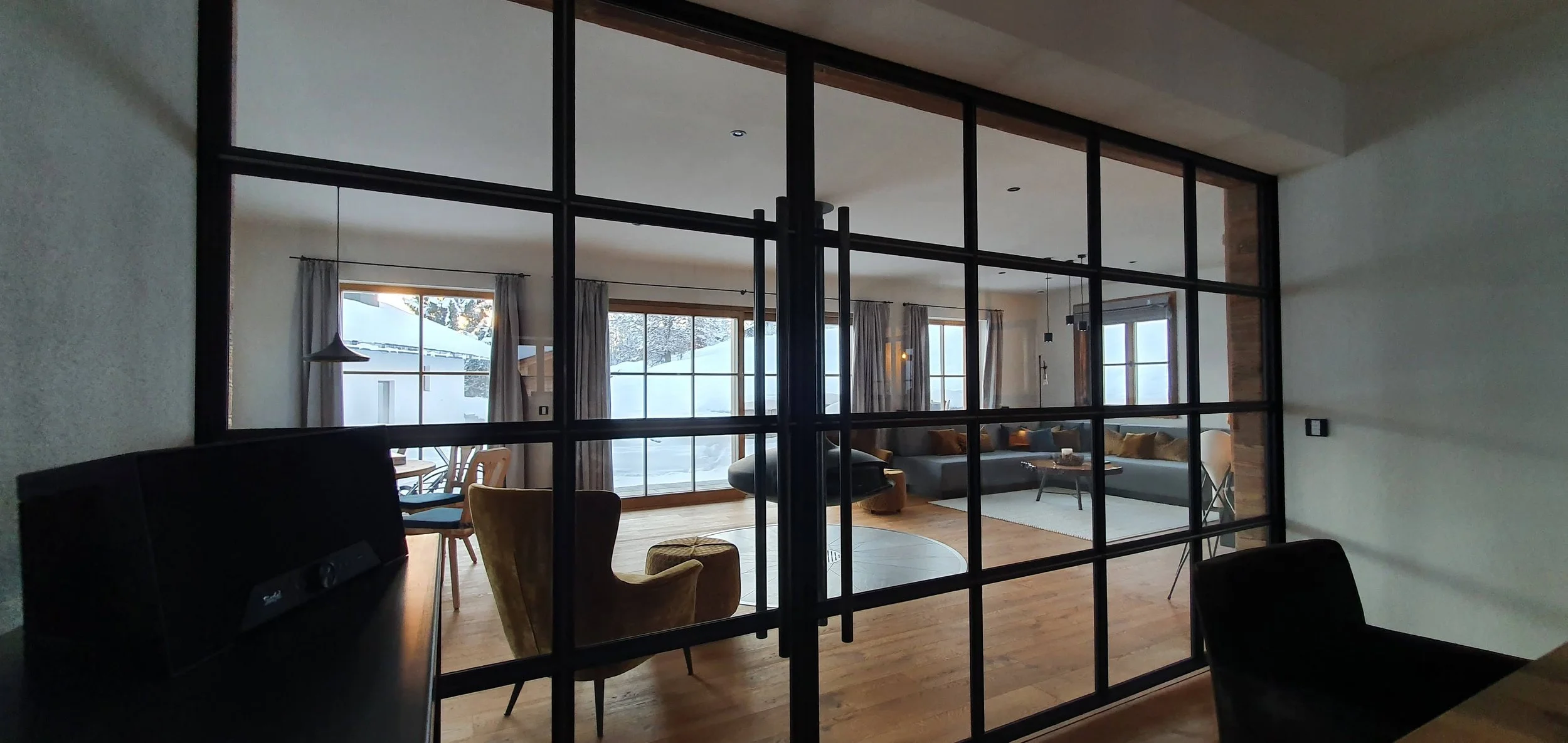 Blick durch eine Glaswand auf ein modernes Wohnzimmer im Winter mit großen Fenstern, Couch, Sessel, Couchtisch und winterlicher Landschaft draußen