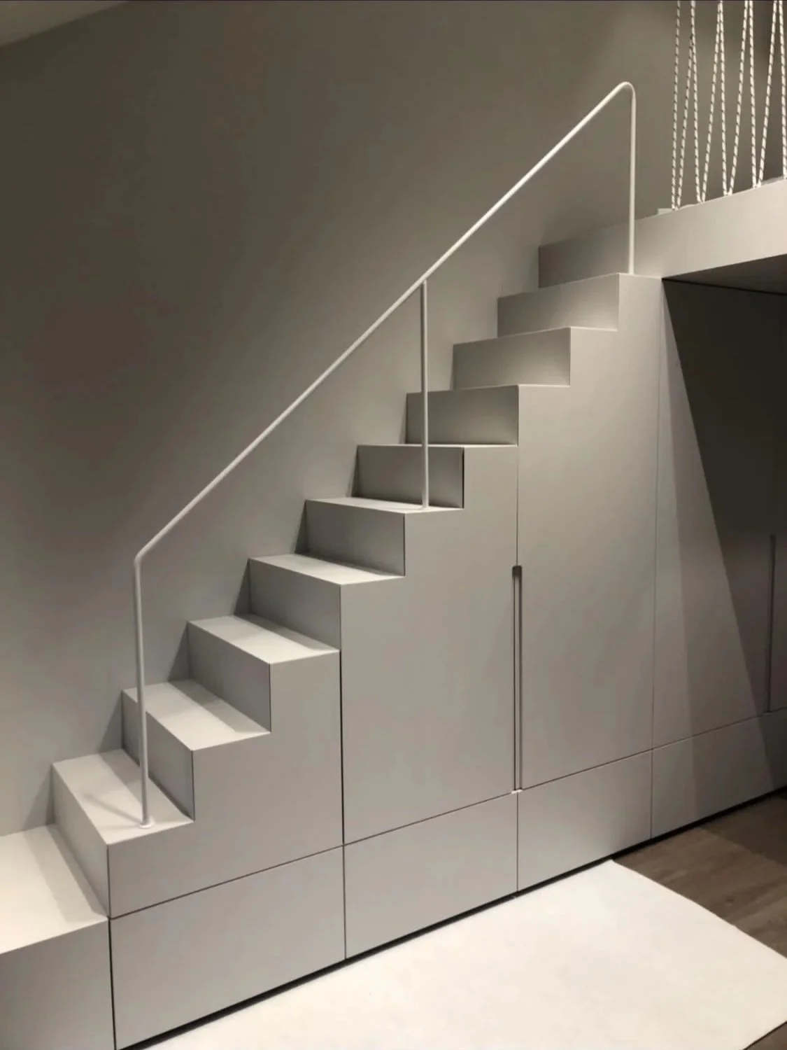 Moderne, weiße Treppe mit abgerundeten Stufen und einem schmalen Handlauf, die in einen Raum mit dunklem Boden führt.