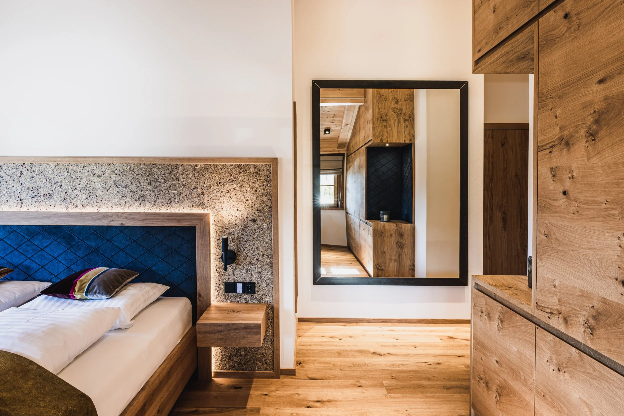 Modernes Schlafzimmer mit Holzdekor, einem großen Spiegel, einem Bett mit blauer gepolsterter Kopfleiste und Nachttisch aus Holz.