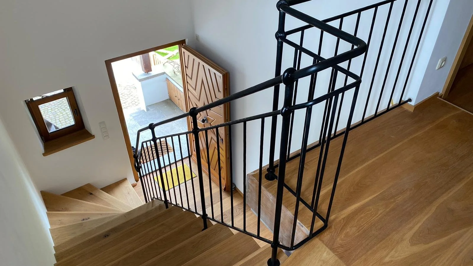 Blick von der oberen Ebene auf den Eingangsbereich mit offenem Holzhaustür, kleinen Fenster und Holztreppe, mit schwarzen Geländer, Holzboden und weißer Wand