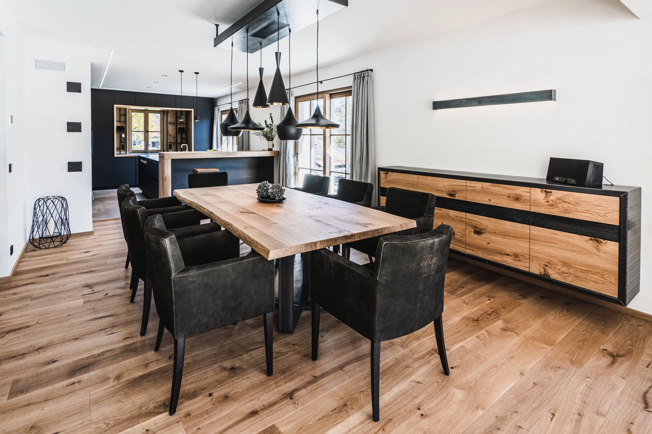 Moderner Essbereich mit Holztisch, schwarzen Ledersesseln und einer Holzsideboard, Fenster mit weißen Vorhängen, schwarze Pendelleuchten über dem Tisch, offener Küchenbereich im Hintergrund.