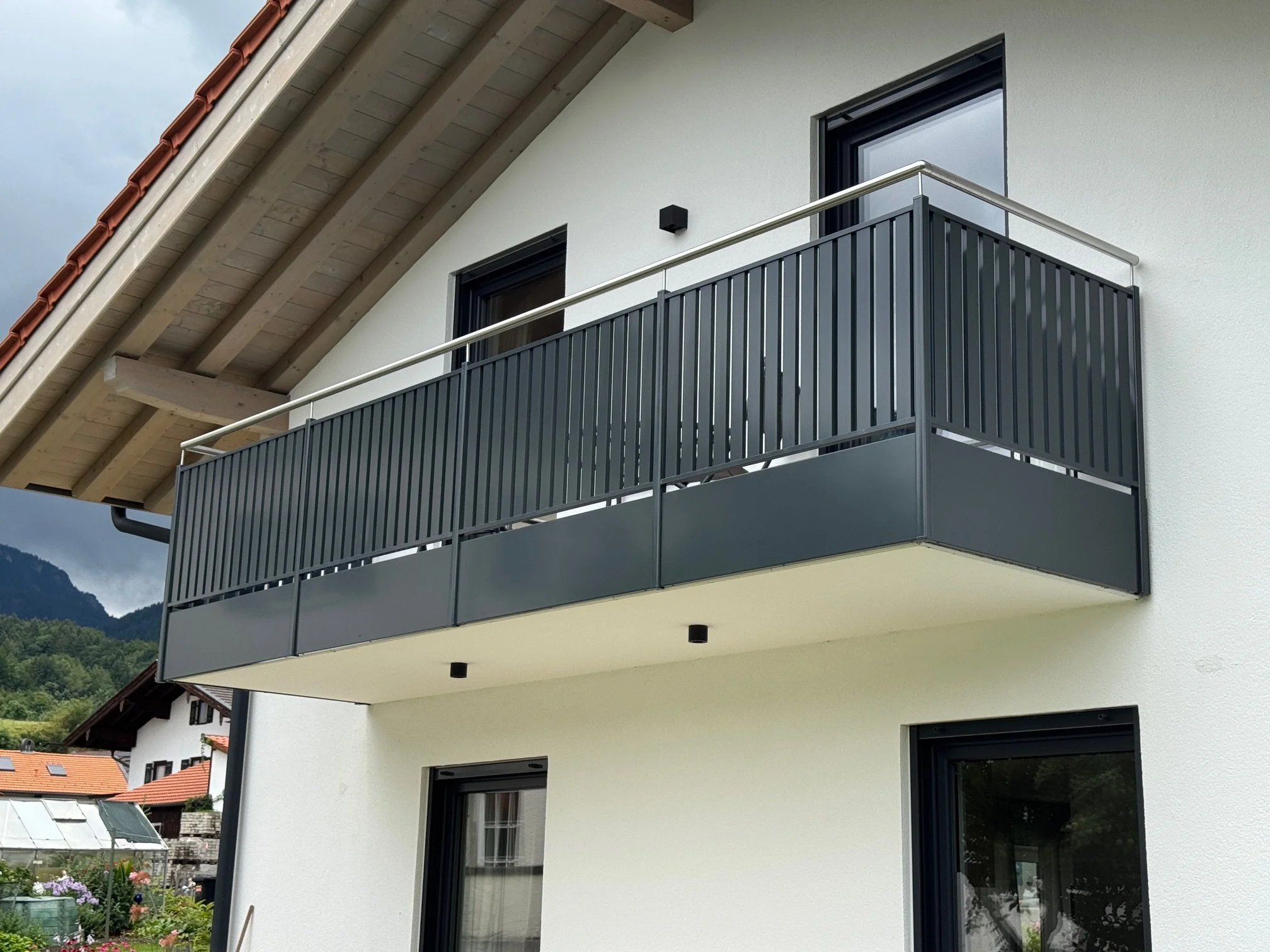 Gerader Balkon mit schwarzen Metallgeländern an einem weißen Haus, mit zwei Fenstern und einem Dach mit roten Ziegeln.