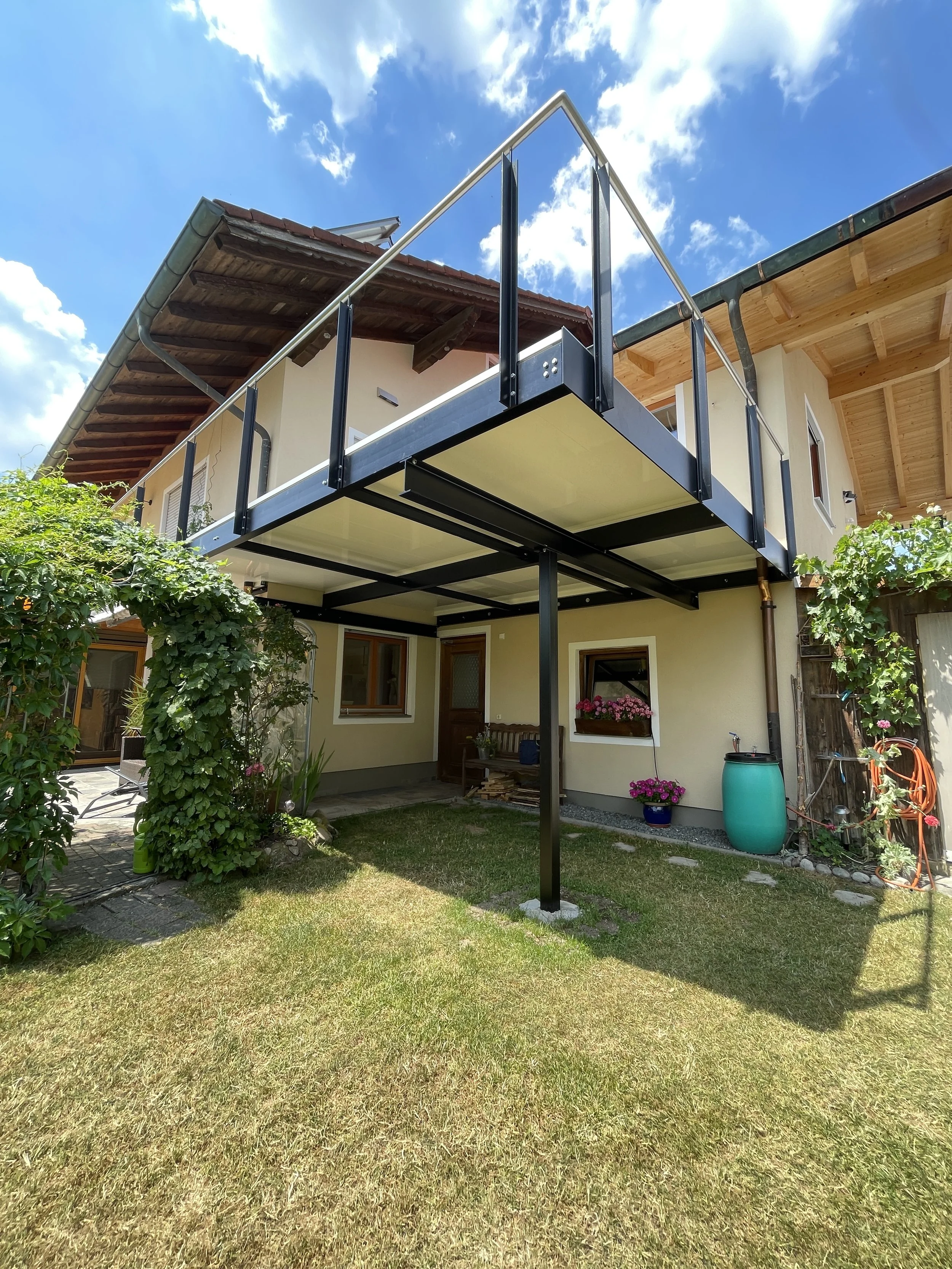 Ein Haus mit Balkon und Garten, der Balkon ist moderne schwarz und beige gestaltet, im Garten Pflanzen und Blumen, an einem sonnigen Tag mit blauen Himmel und Wolken.