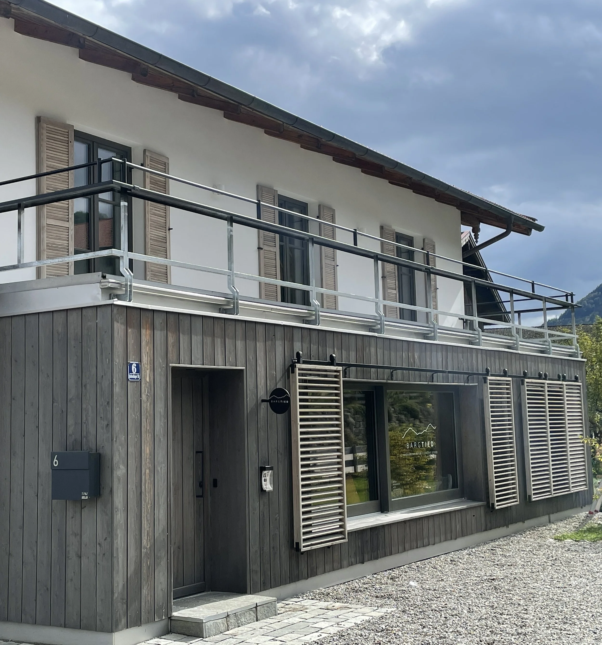 Mehrstöckiges Haus mit Holzwand, grauen Fensterläden und Balkon mit Metallgeländer an der oberen Etage.