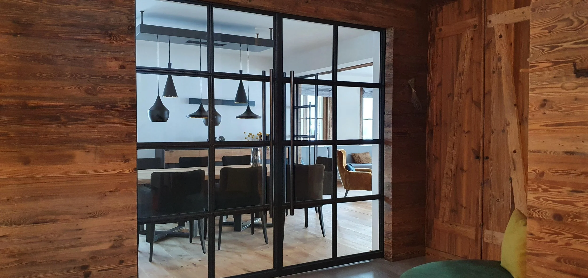 Blick durch eine Glas-Schiebetür in einen modern eingerichteten Essbereich mit schwarzem Tisch und sechs schwarzen Stühlen, sowie einer gelben Sessel im Wohnzimmer, Holzwände und Holzboden