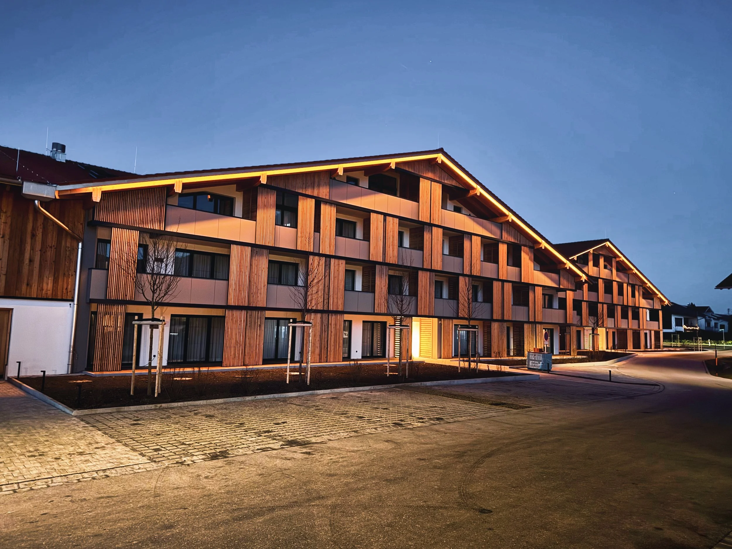 Mehrstöckiges modernes Wohngebäude mit Holzverkleidung bei Nacht, beleuchtet von Außenbeleuchtung, umgeben von einem Parkbereich mit Bäumen und gepflastertem Weg.