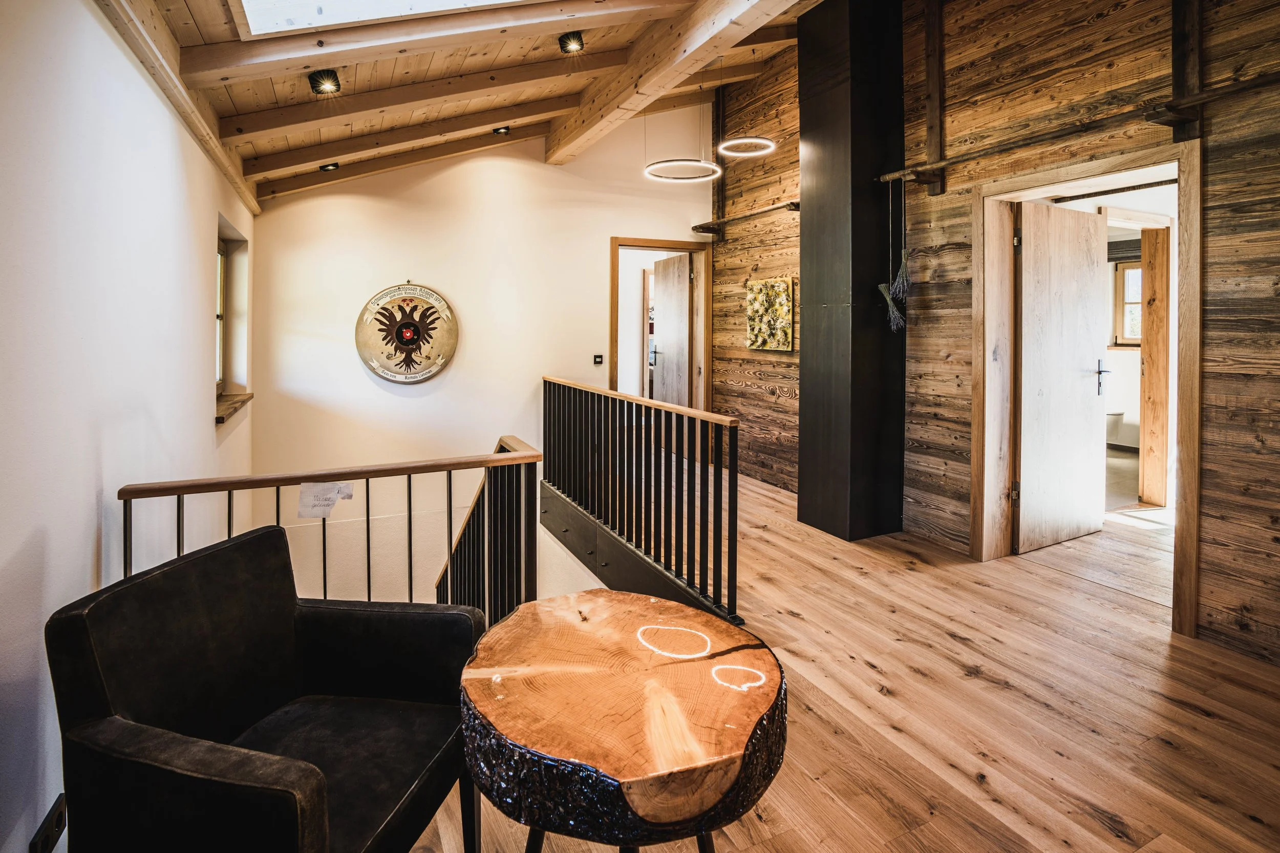 Innenraum eines Hauses mit Holzdecken und Holzwänden, ein schwarzer Sessel, ein runder Holztisch, Kunstwerke an der Wand, offenes Badezimmer mit WC sichtbar.