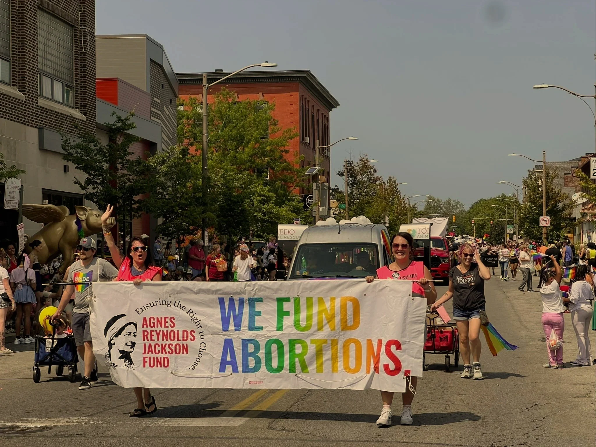 Group of people participating in a pride parade holding a banner that reads 'We Fund Abortions' with a logo of Agnes Reynolds Jackson Fund and rainbow colors, on a city street with onlookers and colorful floats.