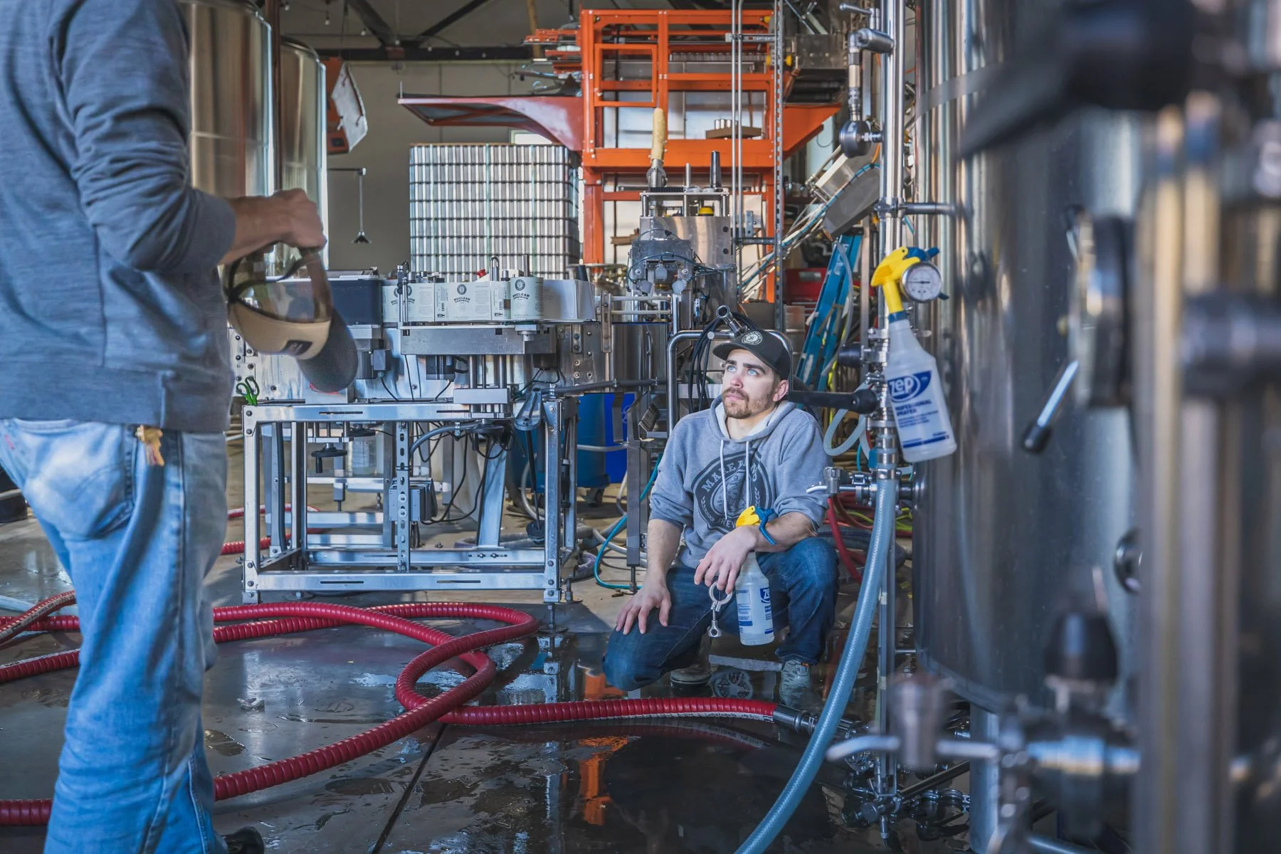 Industrial Photographer Richmond - An Industrial Photographer Richmond captures a man on a brewery floor, spray bottle in hand, amid tanks and industrial equipment.