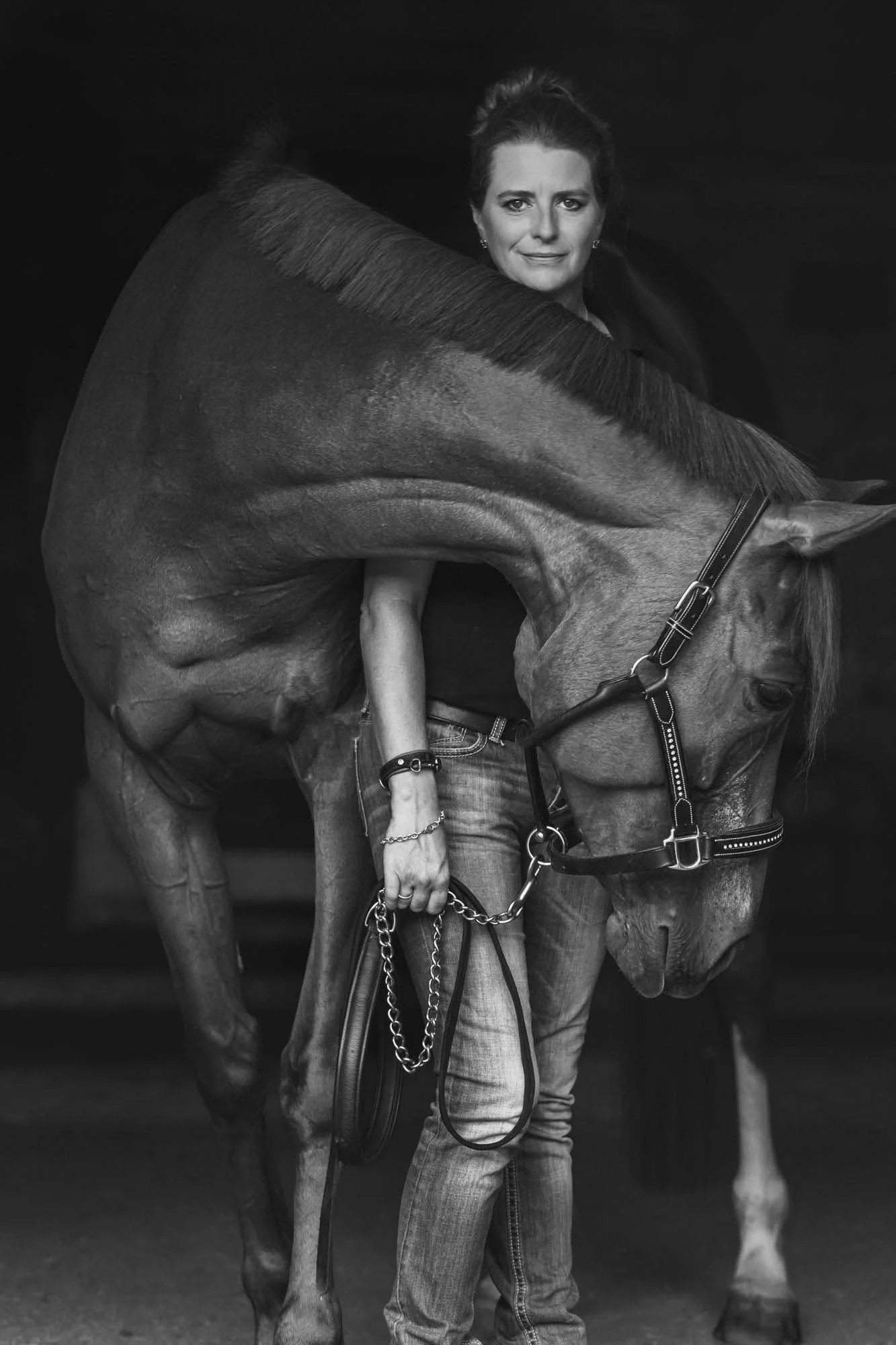 A woman stands calmly as a horse rests its head over her shoulder; both relaxed. Professional brand imagery in black and white.