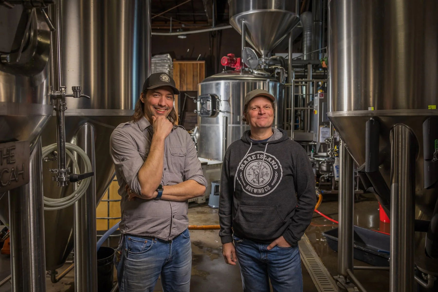 Industrial Photographer Richmond - Two men smiling between large brewing tanks, captured by an Industrial Photographer Richmond amid shiny stainless steel equipment.