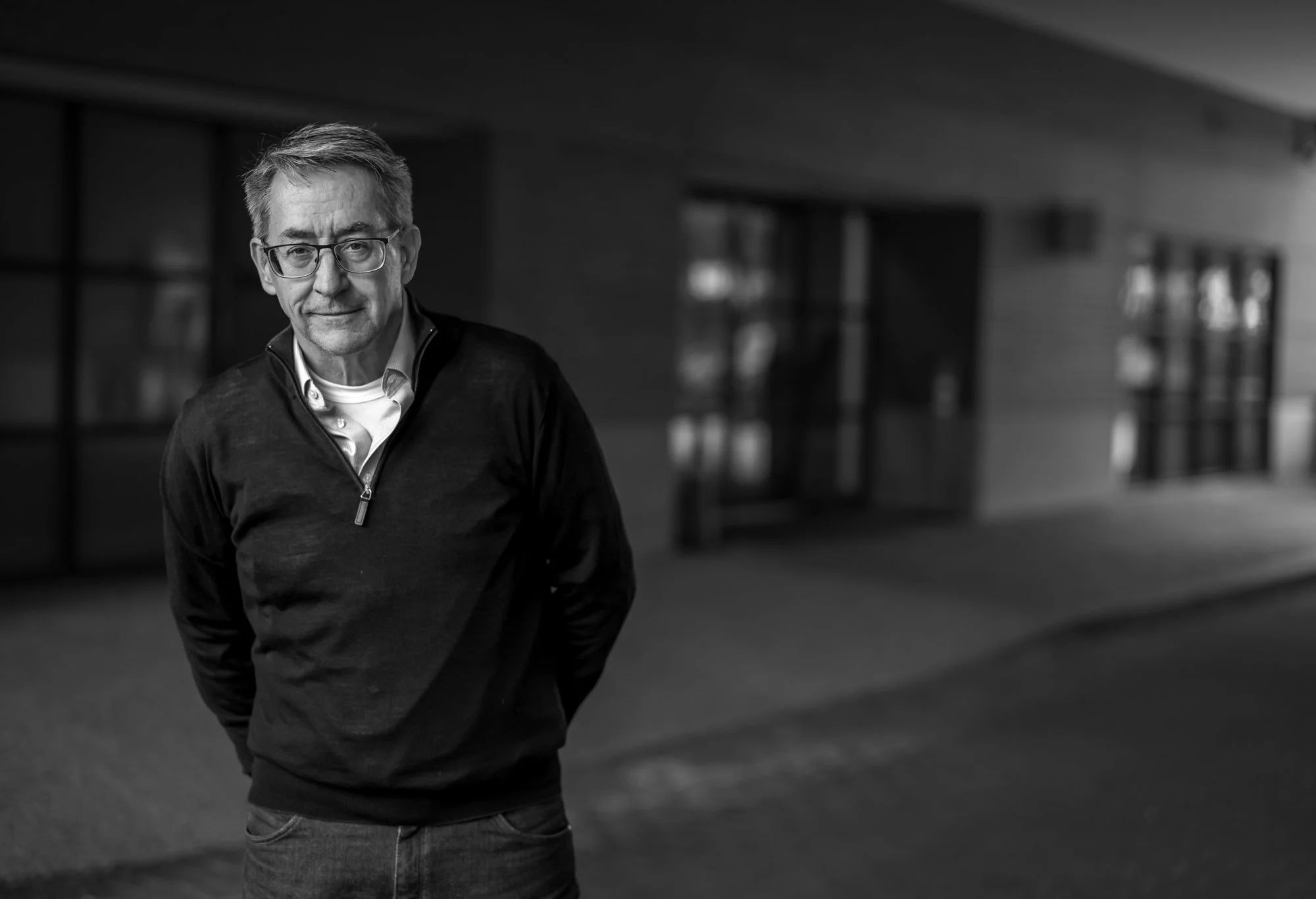 Man in glasses and dark sweater stands outside a building, slight smile; blurred background