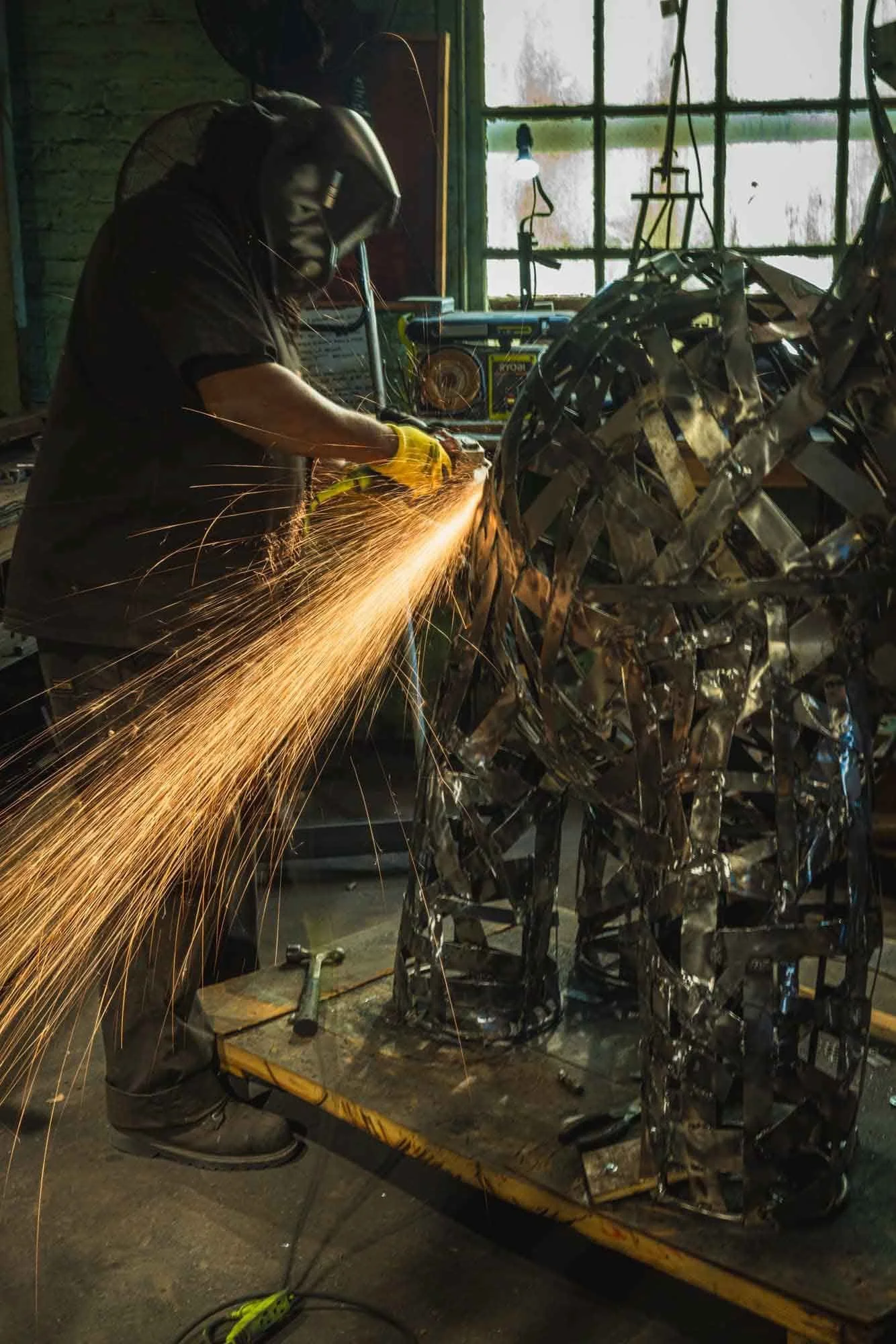 A person in a welding mask uses a grinder on a metal sculpture